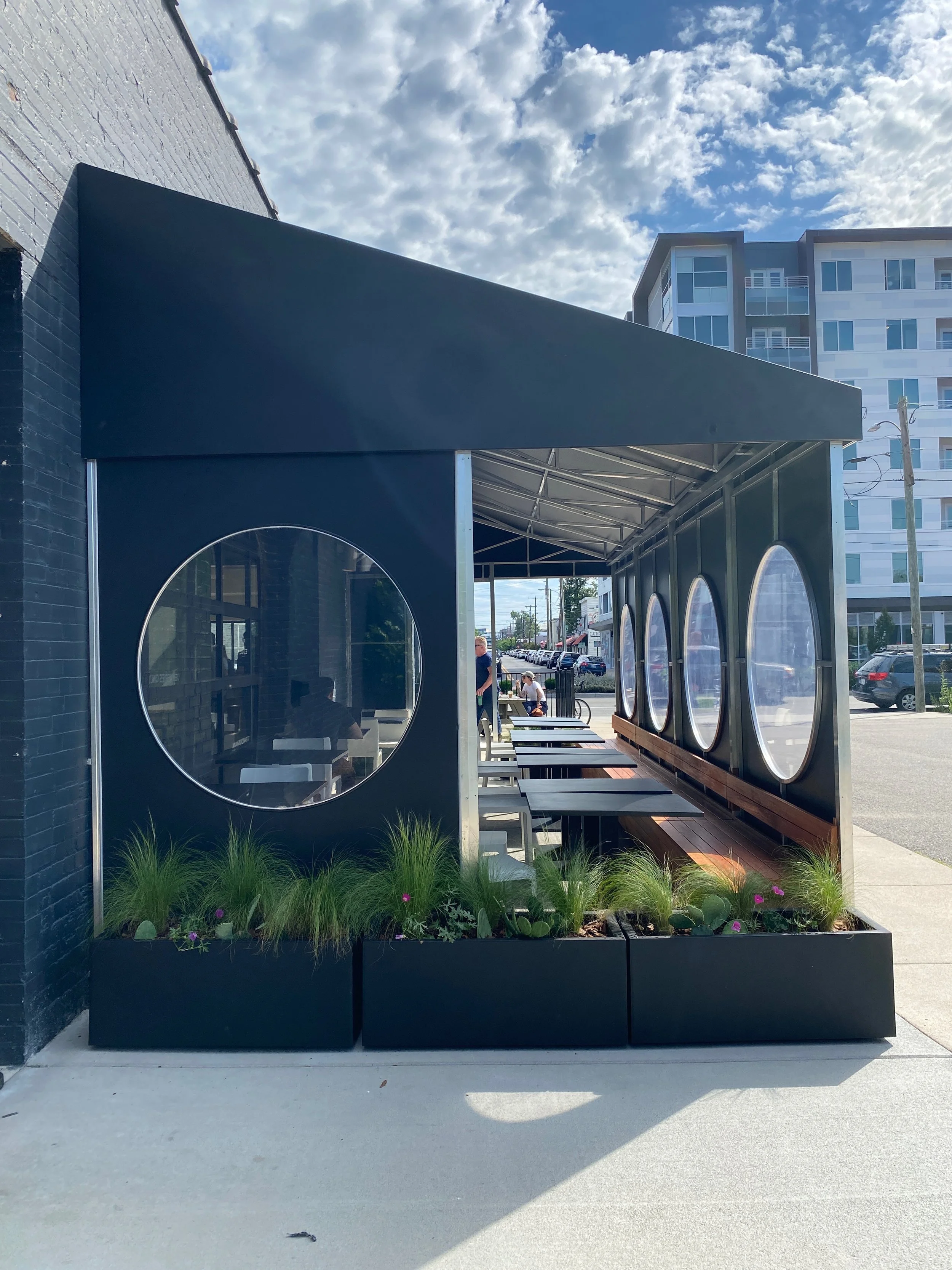 Outdoor seating area with iron tables and benches under a canopy, with circular windows in the canopy walls, and surrounding planters with green grass and purple flowers. In the background, a street with parked cars and a multi-story building is visi