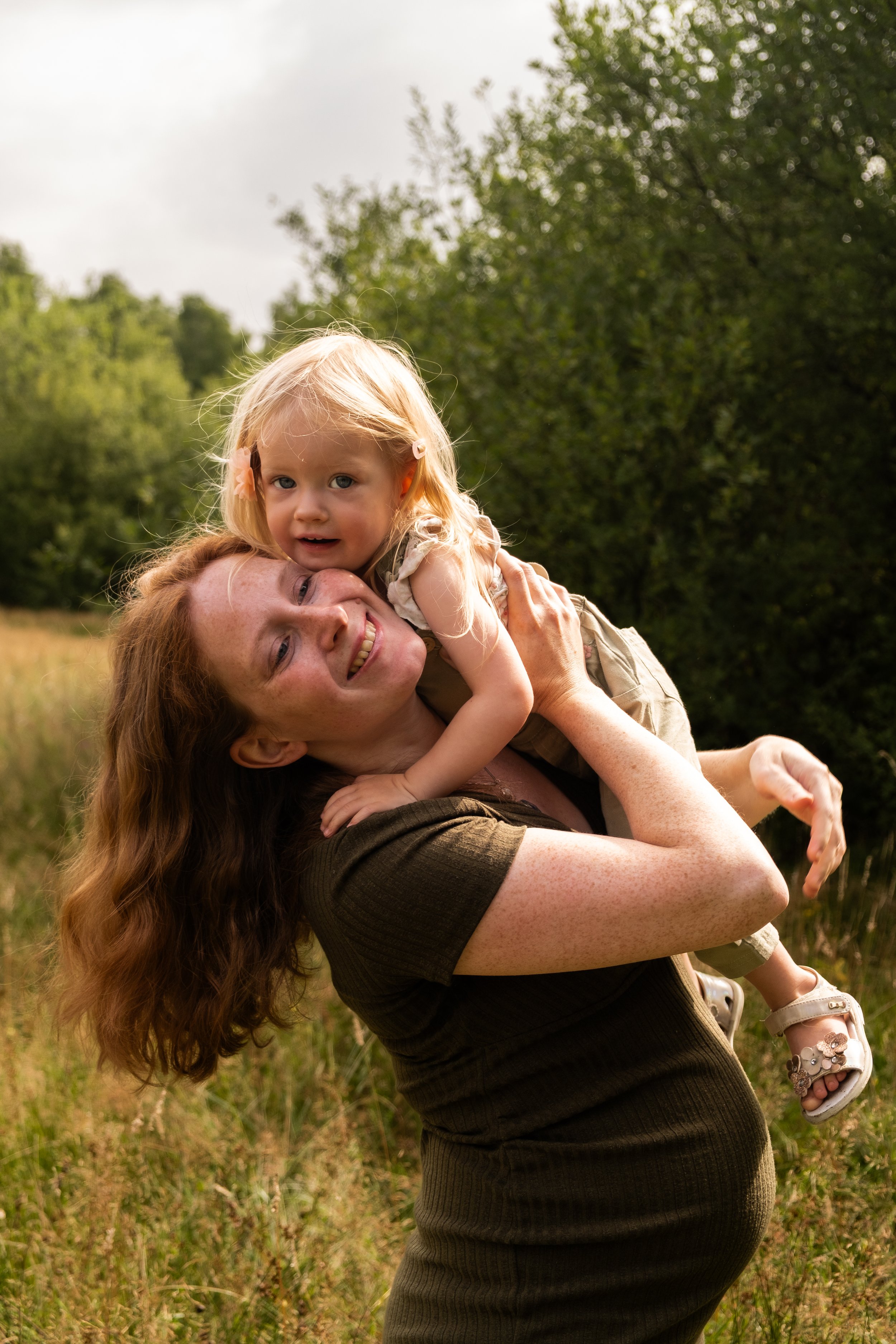Vrouw met lang krullend haar die een klein meisje op haar schouders vasthoudt in een veld met gras en bomen op de achtergrond. Ze glimlachen allebei naar de camera bij zonnig weer.
