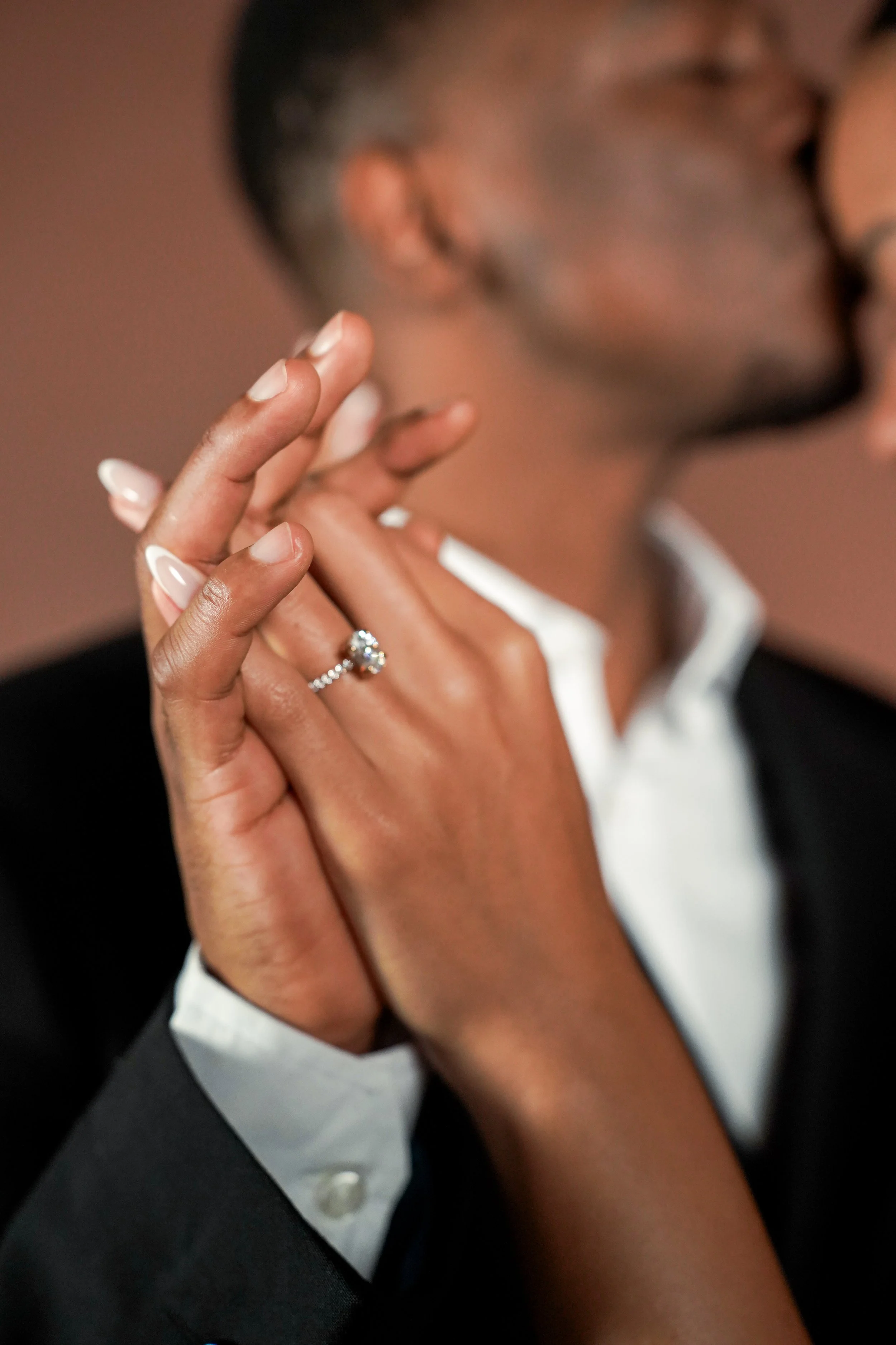 Close-up van een hand met een diamanten verlovingsring, de persoon draagt een zwarte pak en een witte overhemd, de achtergrond is vaag.
