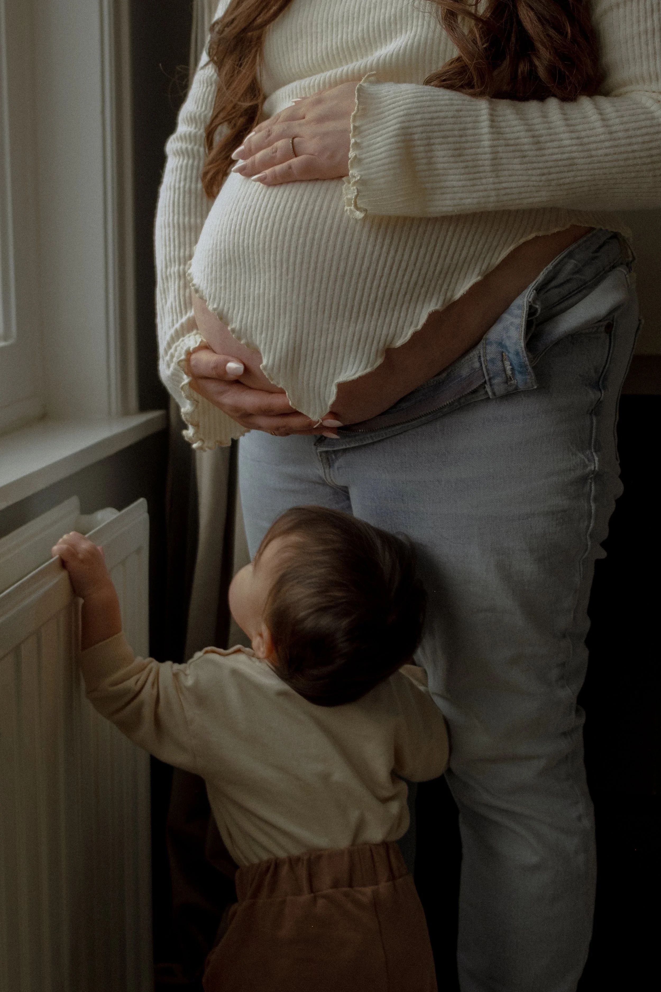 Een vrouw houdt haar zwangere buik vast terwijl een kind omhoog kijkt en het voetje op de radiator legt.