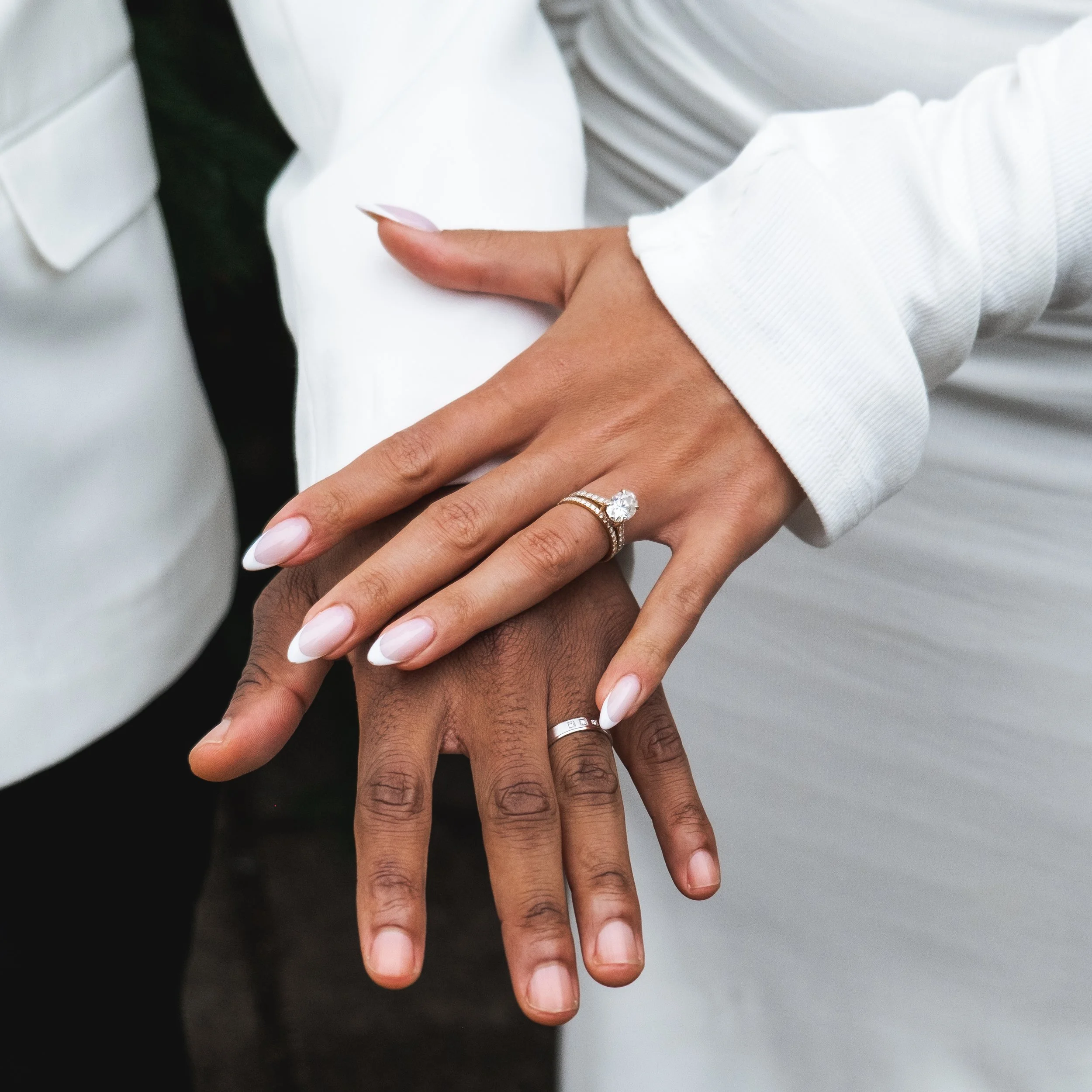 Twee mensen die elkaars handen vasthouden, één persoon met een diamanten verlovingsring en een ring. Ze dragen witte en zwarte kleding.