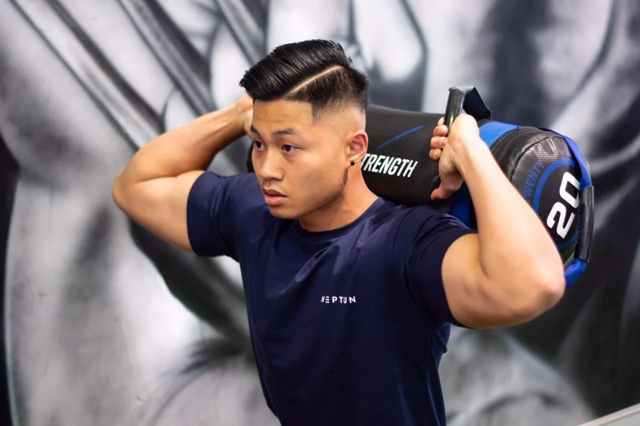 A man in a navy blue shirt carrying a black and blue bag on his shoulders at the gym, with a muscular build and short black hair.