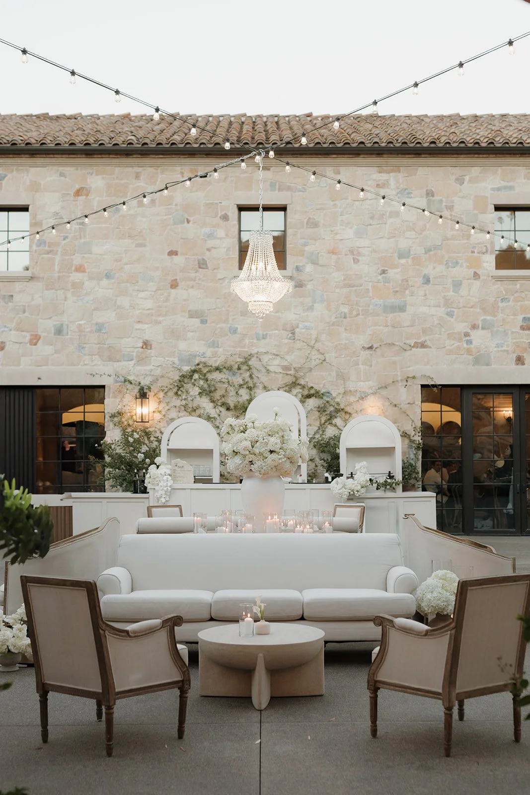 Elegant outdoor lounge setup with white sofa, chairs, and table in front of a stone wall decorated with green vines, surrounded by floral arrangements and candles, under string lights and a chandelier.