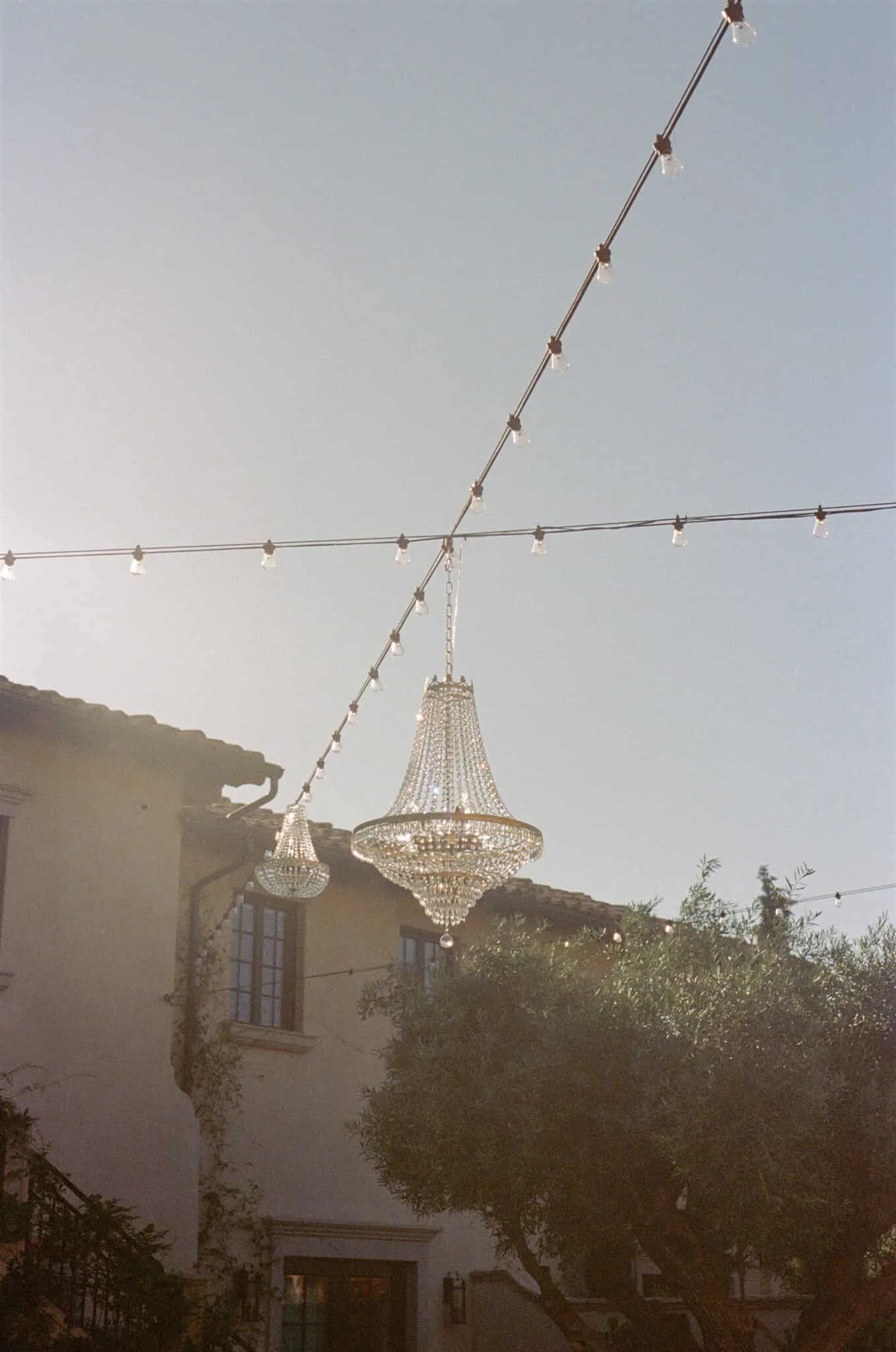 Outdoor scene with string lights and two chandeliers hanging in the sky, with a building and trees in the background.