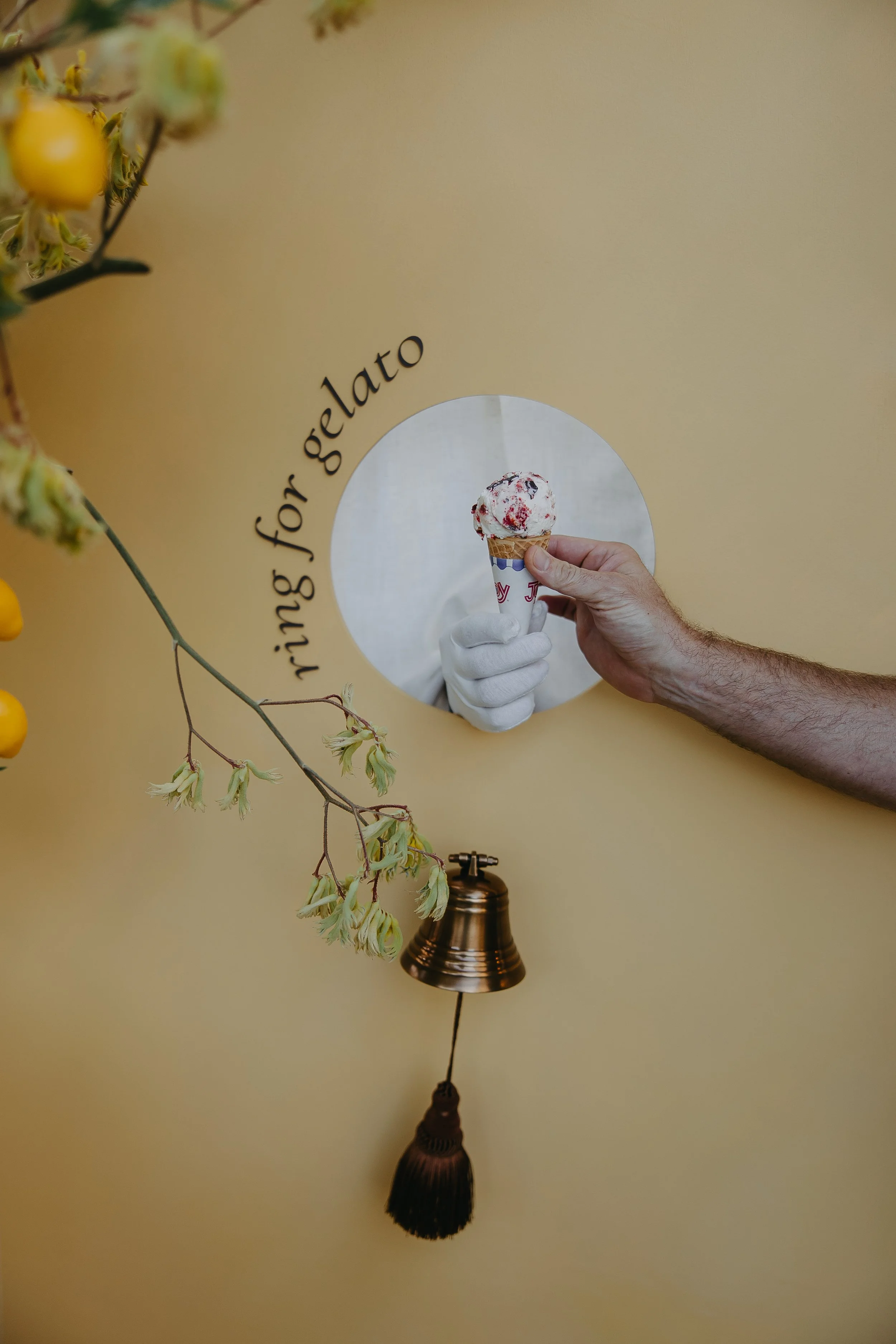 Close-up of a person's arm and hand, wearing a white glove, holding a scoop of strawberry ice cream in a cone through a circular hole in a yellow wall. The wall has the text 'rising for gelato' beside the hole, and a brass bell with a tassel hangs below. There are some yellow flowers in the foreground.