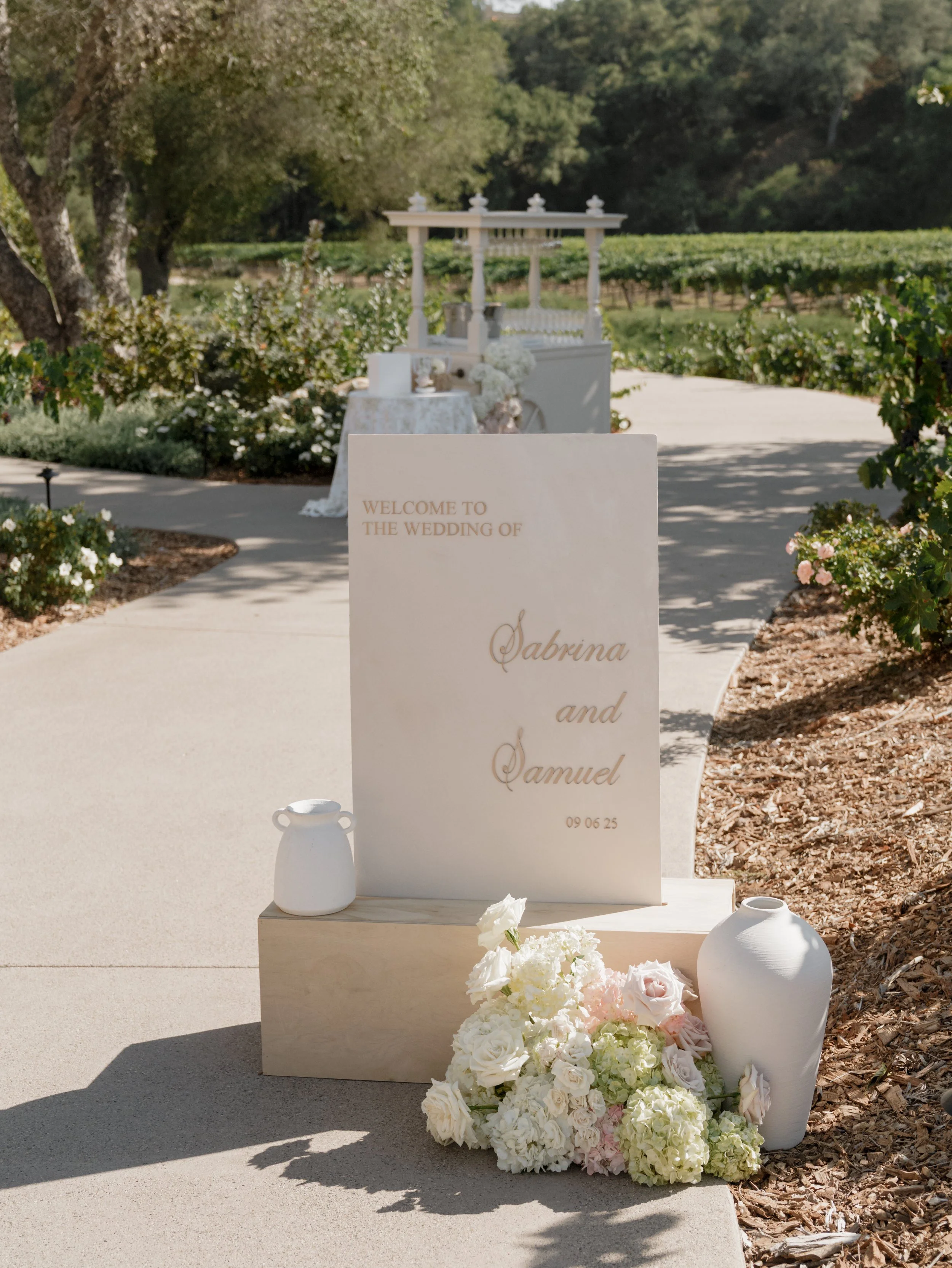 Wedding welcome sign for Sabrina and Samuel, date 09/06/25, with white vases and bouquets on a sidewalk outside a decorated outdoor venue surrounded by greenery.