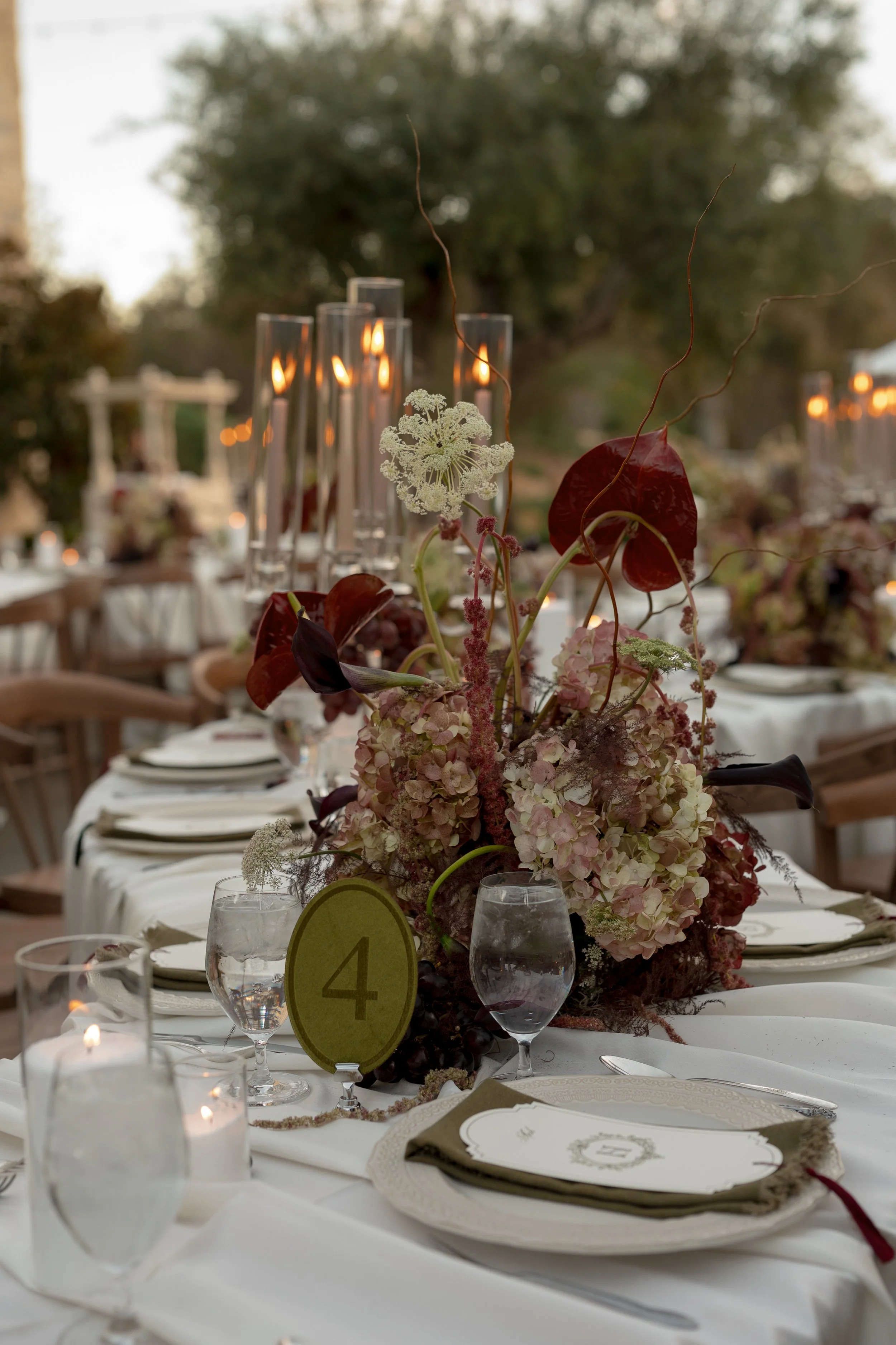 Elegant outdoor dining table set for a wedding or formal event, decorated with a floral centerpiece in shades of pink and burgundy, lit candles, glassware, and table settings, with trees and string lights in the background.