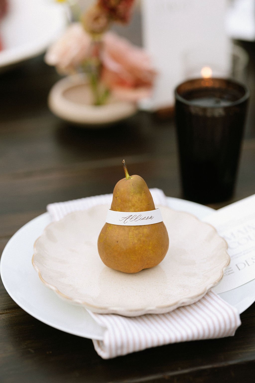 A pear with a white ribbon labeled 'Allison' rests on a beige ceramic plate on a white charger, set on a striped napkin at a table. In the background, a black cup and a blurred flower arrangement are visible.