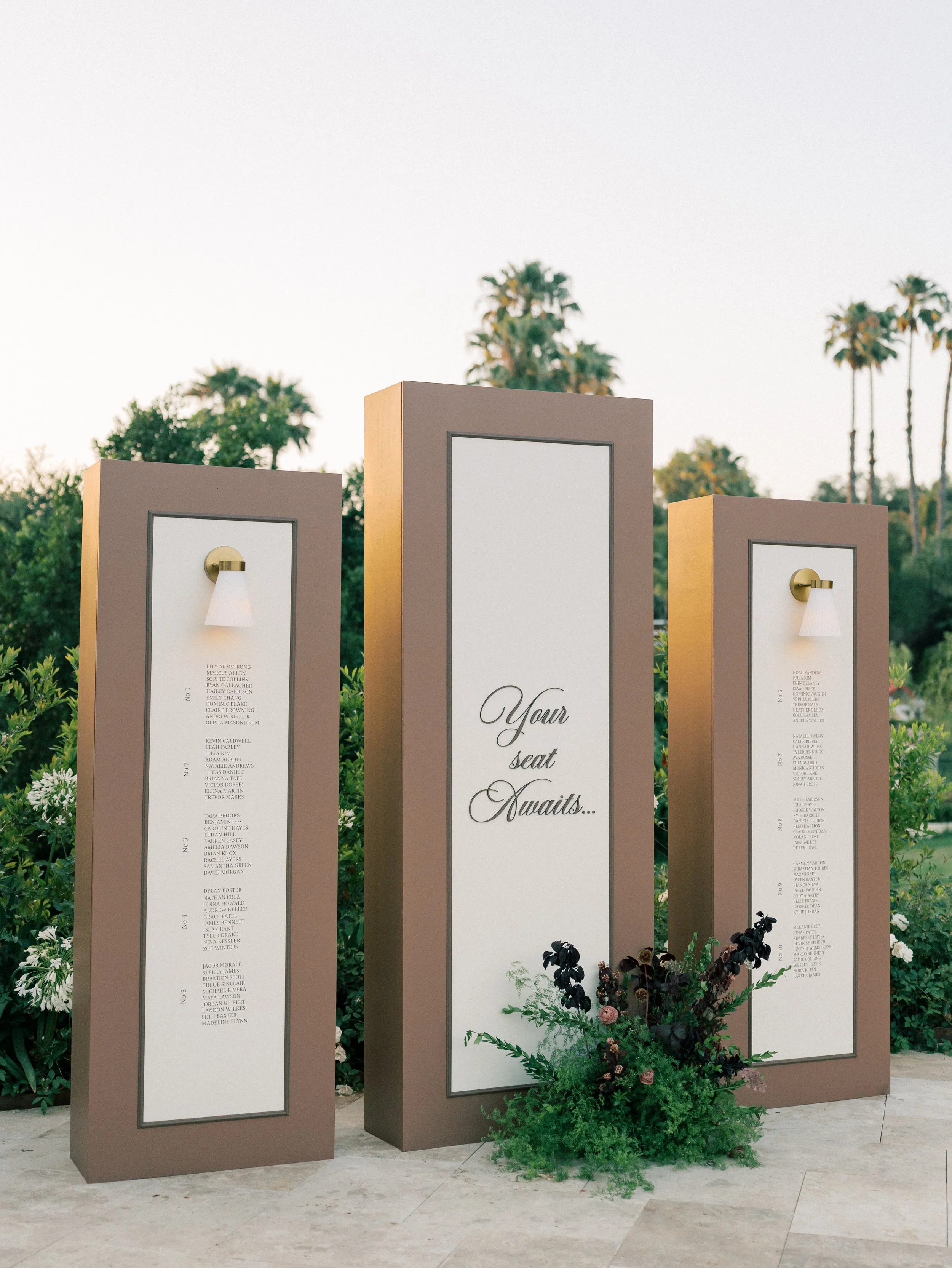 Three standing emoji boards, with the middle one displaying the message 'Your seat awaits...' and the two flanking boards showing rows of names, with small wall lamps on top of the side boards, and a flower arrangement at the base, set outdoors against a backdrop of trees and clear sky.