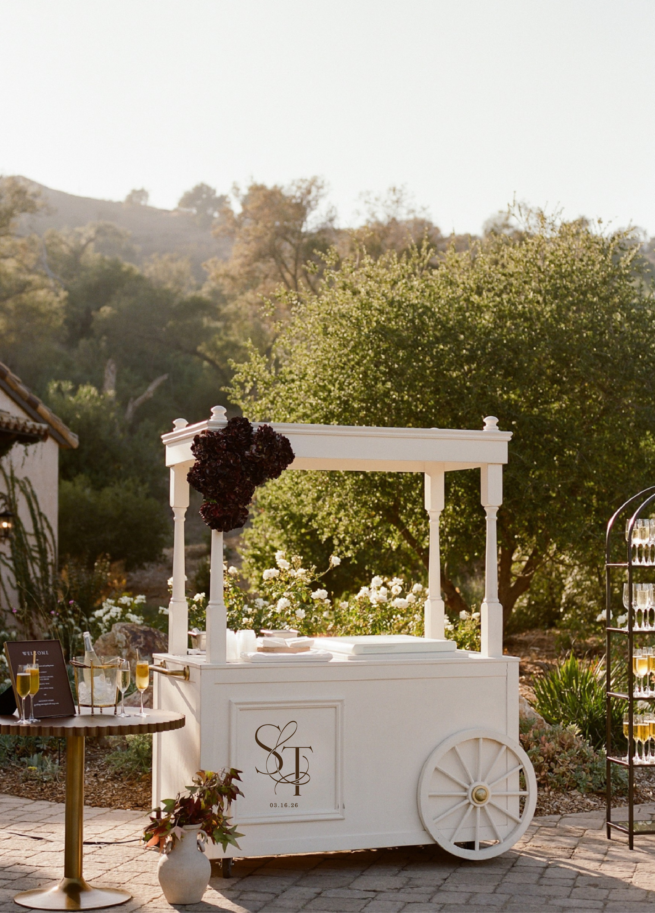 A white wedding cart decorated with dark flowers, with a monogram and date, surrounded by gold champagne glasses, a welcome sign, and outdoor garden setting with trees and flowers.