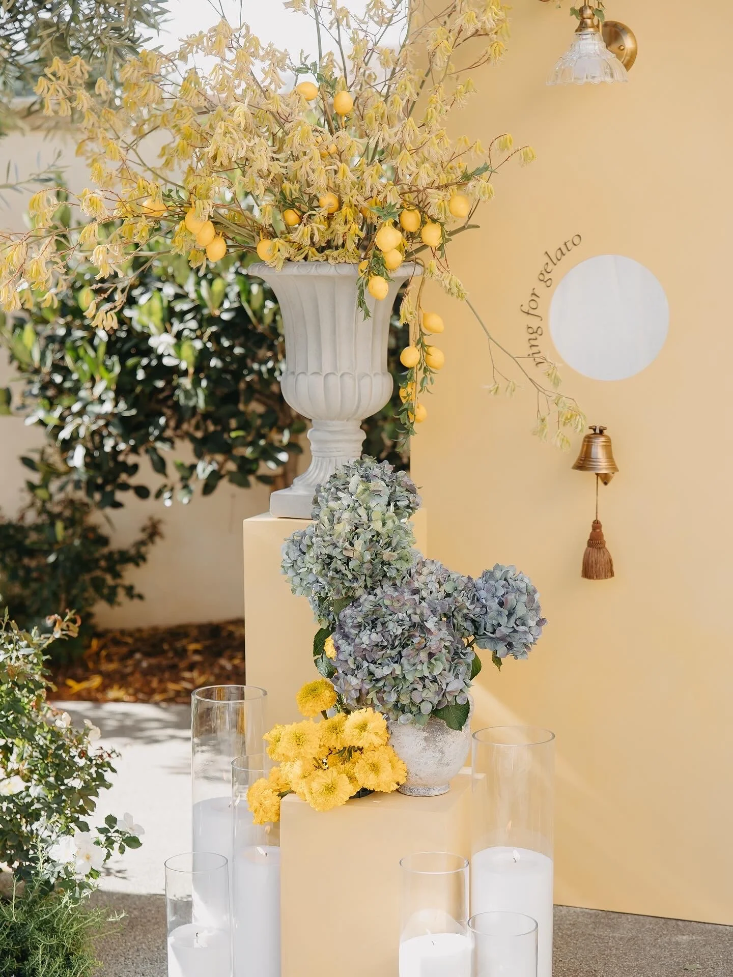 Decorative floral arrangement with yellow, purple, and white flowers in white vases, surrounded by candles and greenery, set against a pastel yellow backdrop.