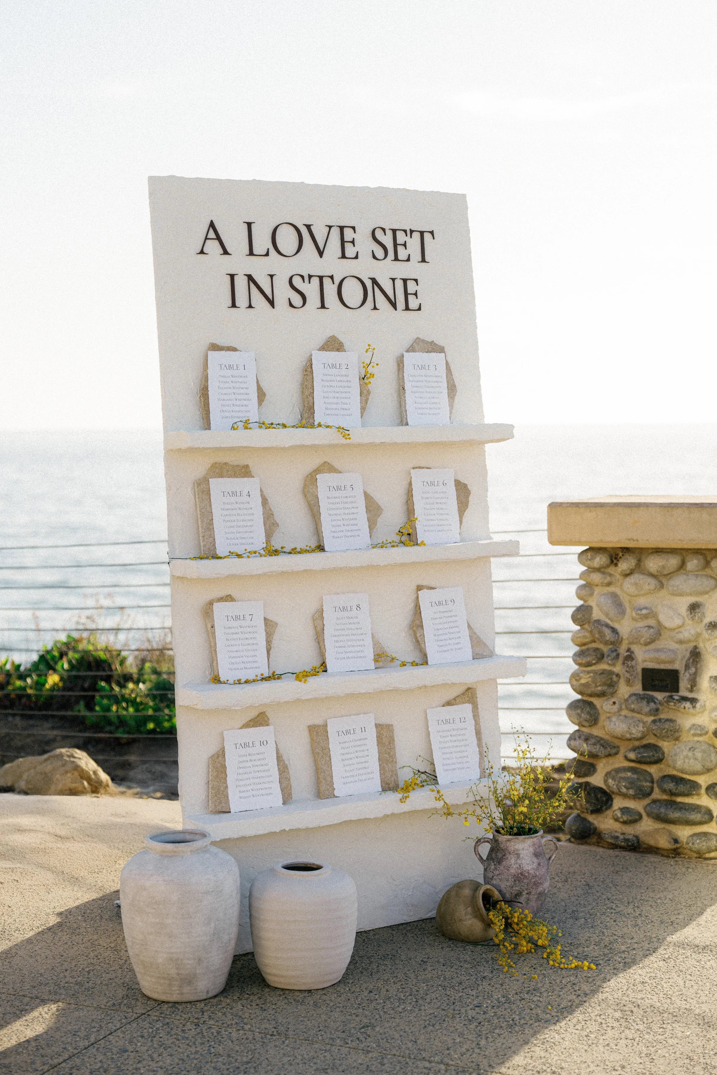 Seating chart display titled 'A Love Set in Stone' with numbered table cards, decorative vases, and flowers, by the ocean.