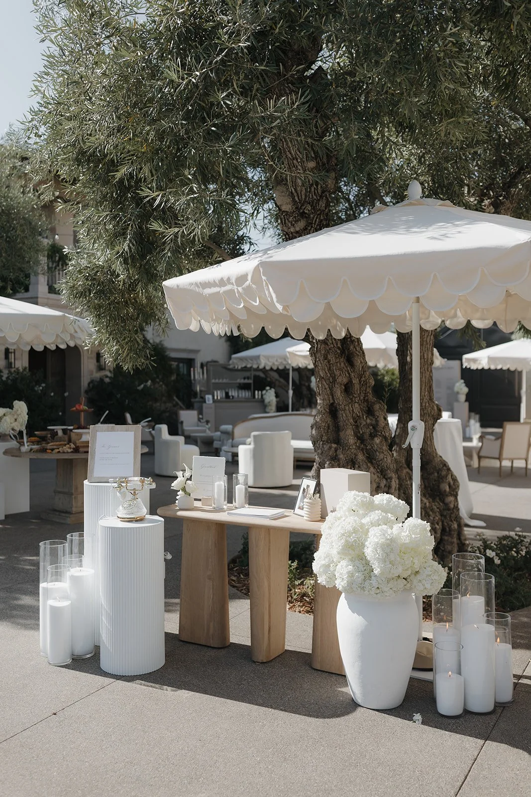Outdoor wedding or event setup with white umbrellas, large white flower arrangements, candles in glass holders, and a wooden table with decorative items under a large tree.