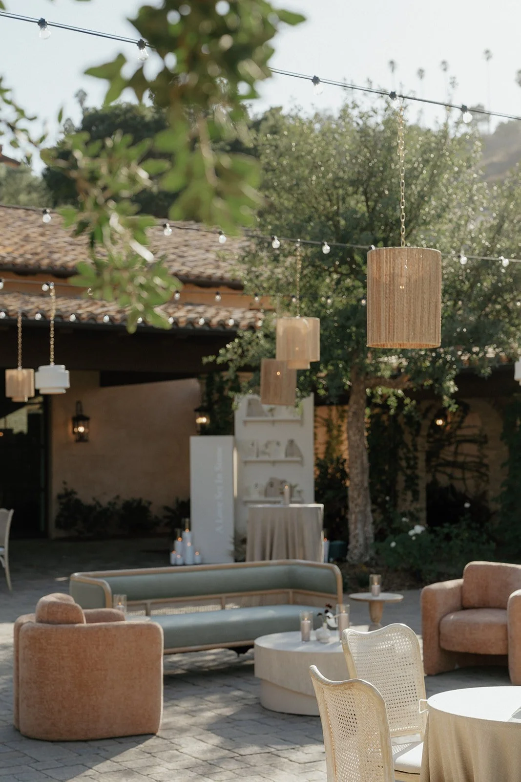 An outdoor patio area with hanging wooden lanterns, string lights, cushioned seating, small tables, trees, and a building with a tiled roof in the background.