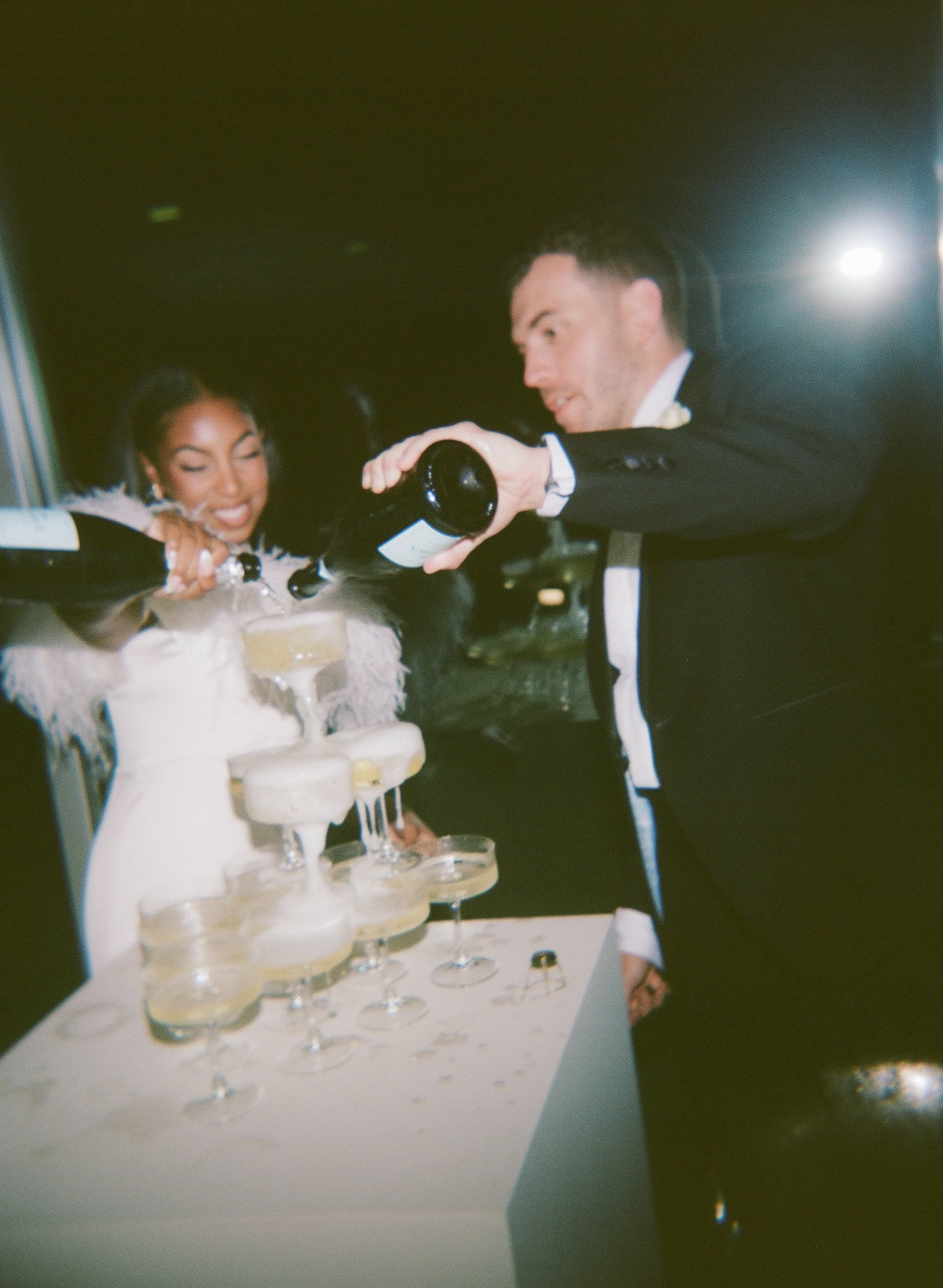 Two people, a woman in a white dress and a man in a black tuxedo, pour champagne into a pyramid of glasses at a celebration event.