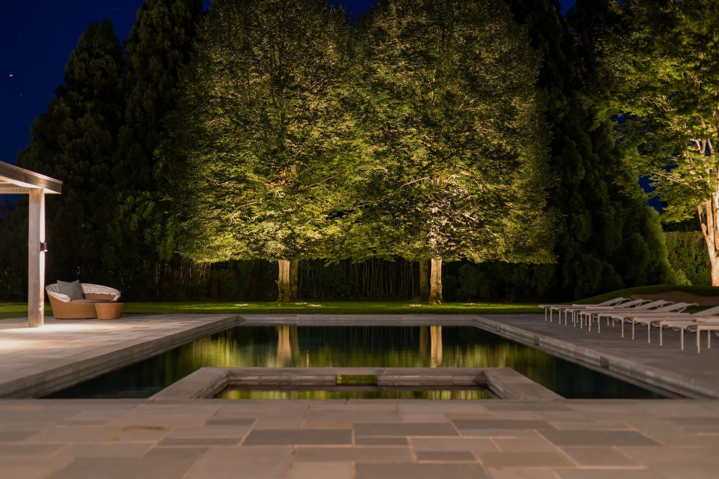 Nighttime scene of a backyard with a pool, surrounded by lawn chairs, a lounge chair with cushions, and large illuminated trees in the background.