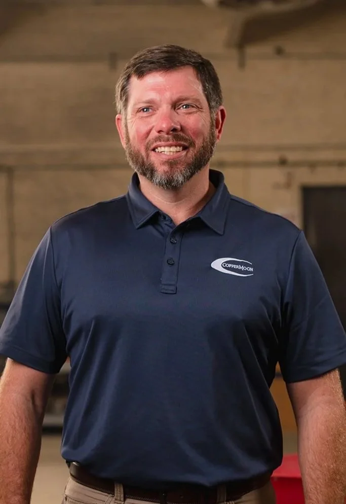 A man with a beard and short hair wearing a navy polo shirt with a 'CopperMoon' logo, standing in a wooden workshop or industrial space.