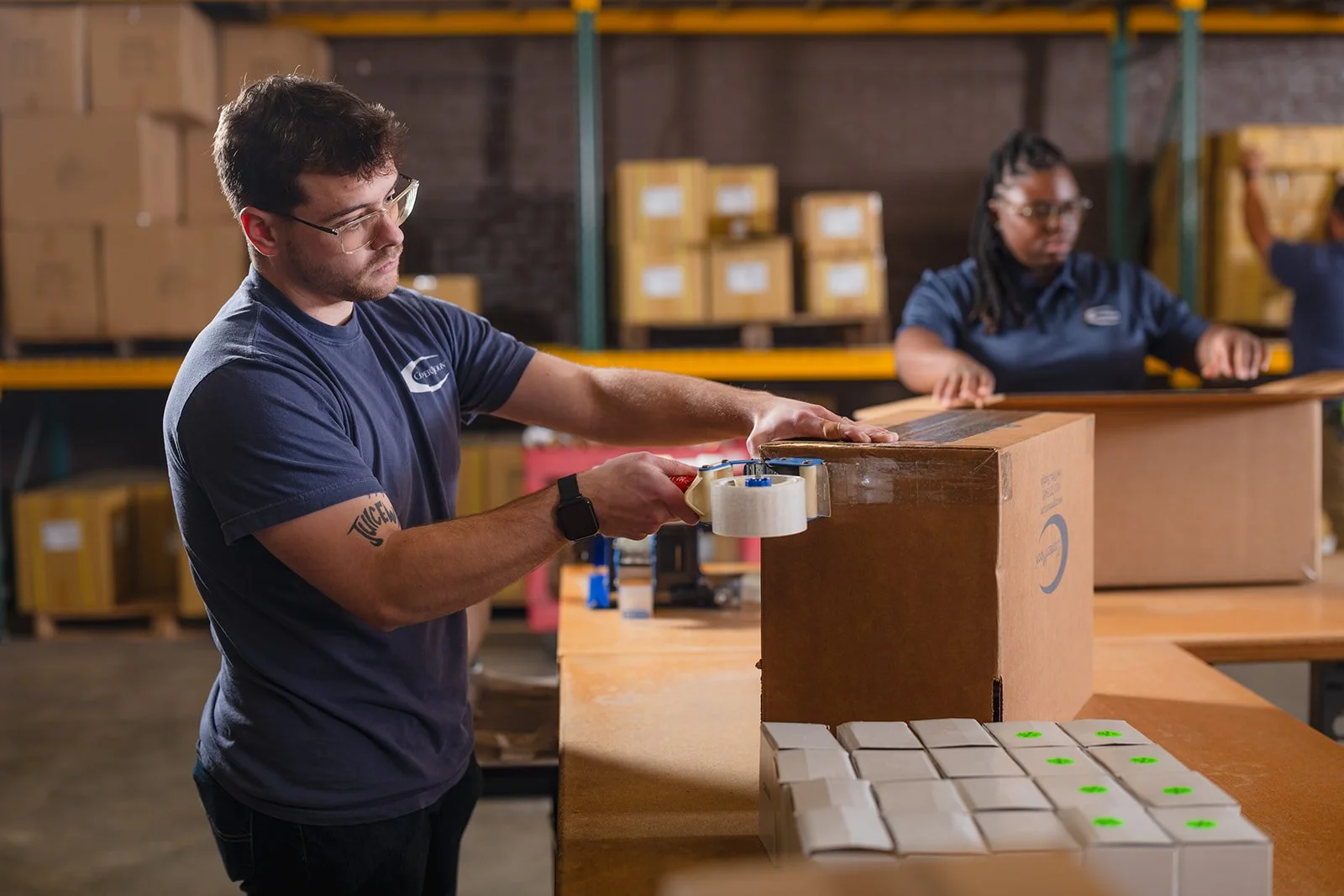 A man is sealing a cardboard box with packing tape in a warehouse, with two women working in the background.