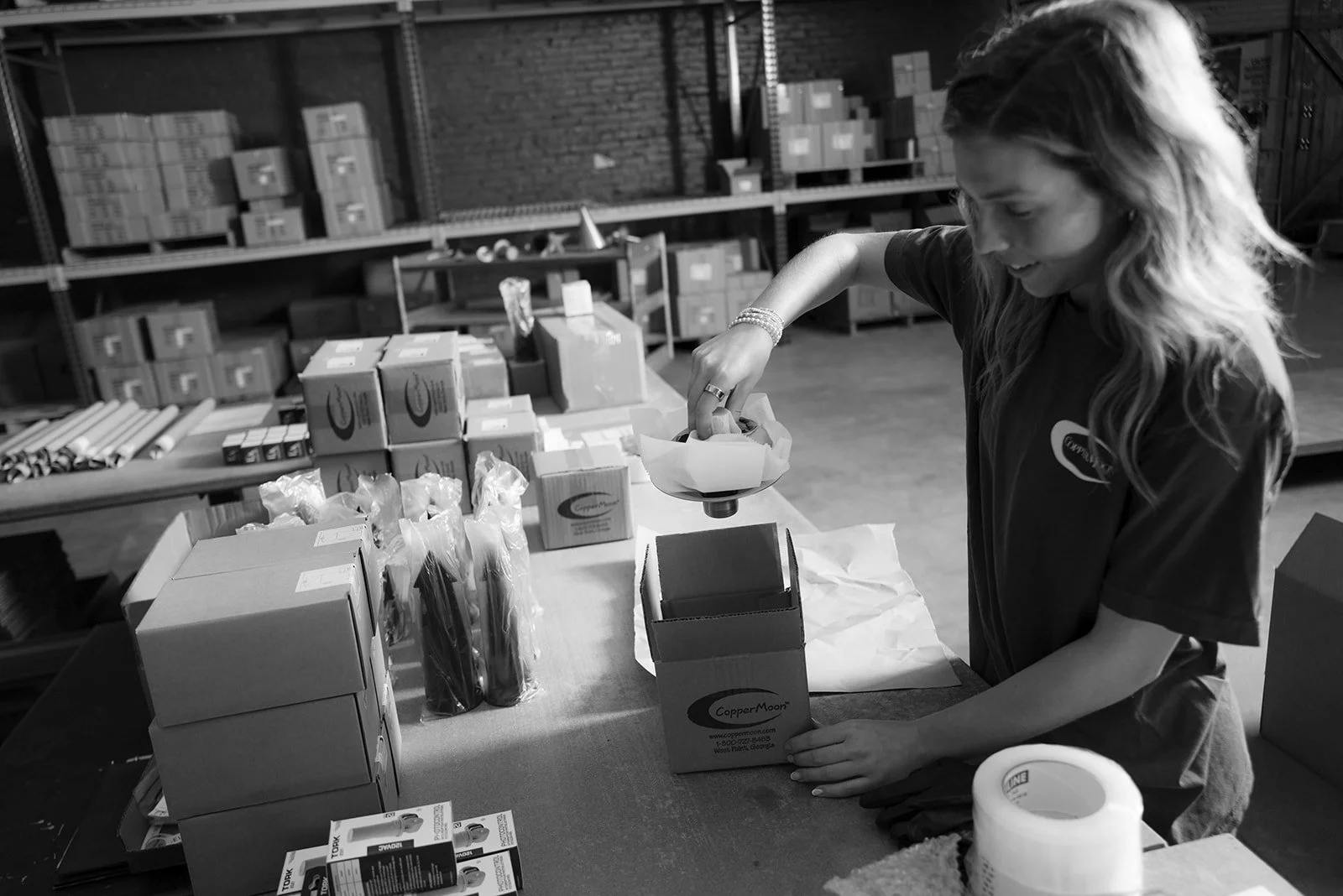 A woman working in a warehouse, packing boxes on a table surrounded by supplies and storage shelves filled with boxes.