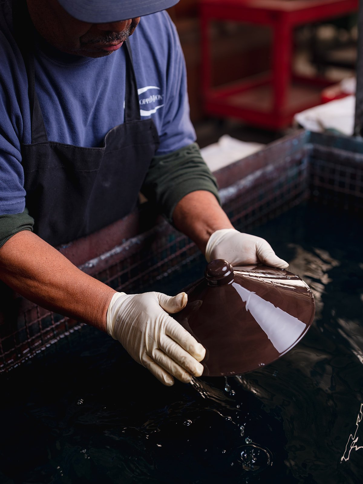 A person wearing gloves and a blue shirt holding a shiny, glazed piece of pottery over a water-filled container.