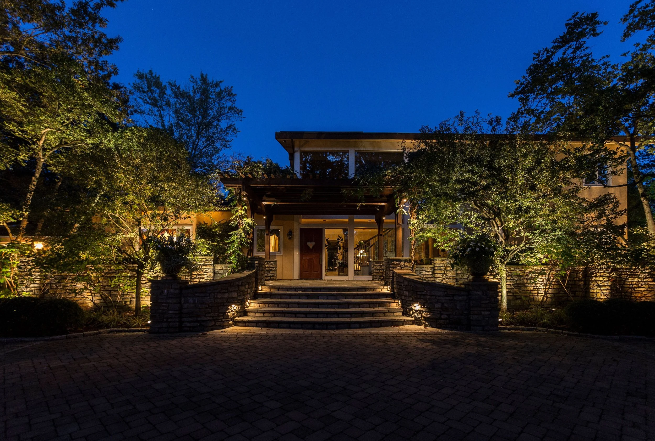 A house illuminated at night, with stairs leading up to the front door, surrounded by trees and garden lighting.