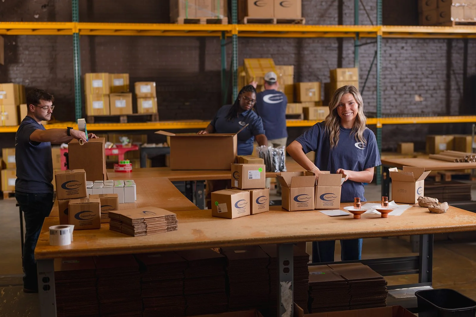 People working in a warehouse packing boxes at a large wooden table, with shelves stocked with boxes in the background.