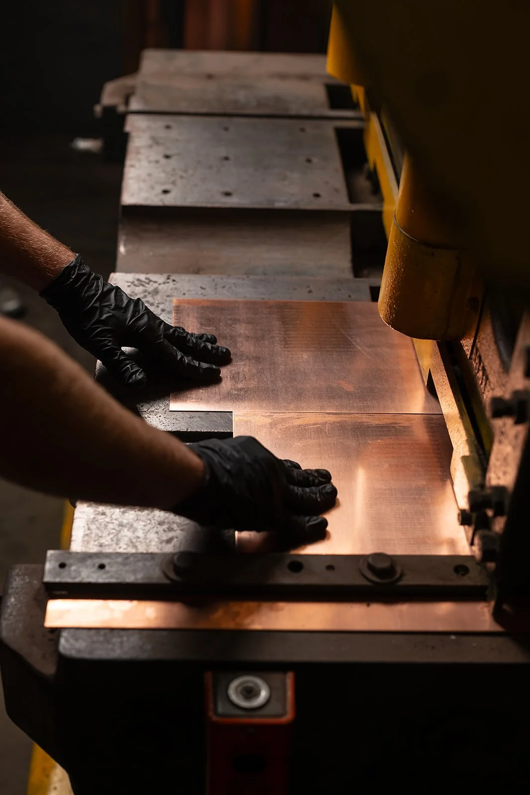 A person wearing black gloves working with a copper sheet on a machine in a workshop.