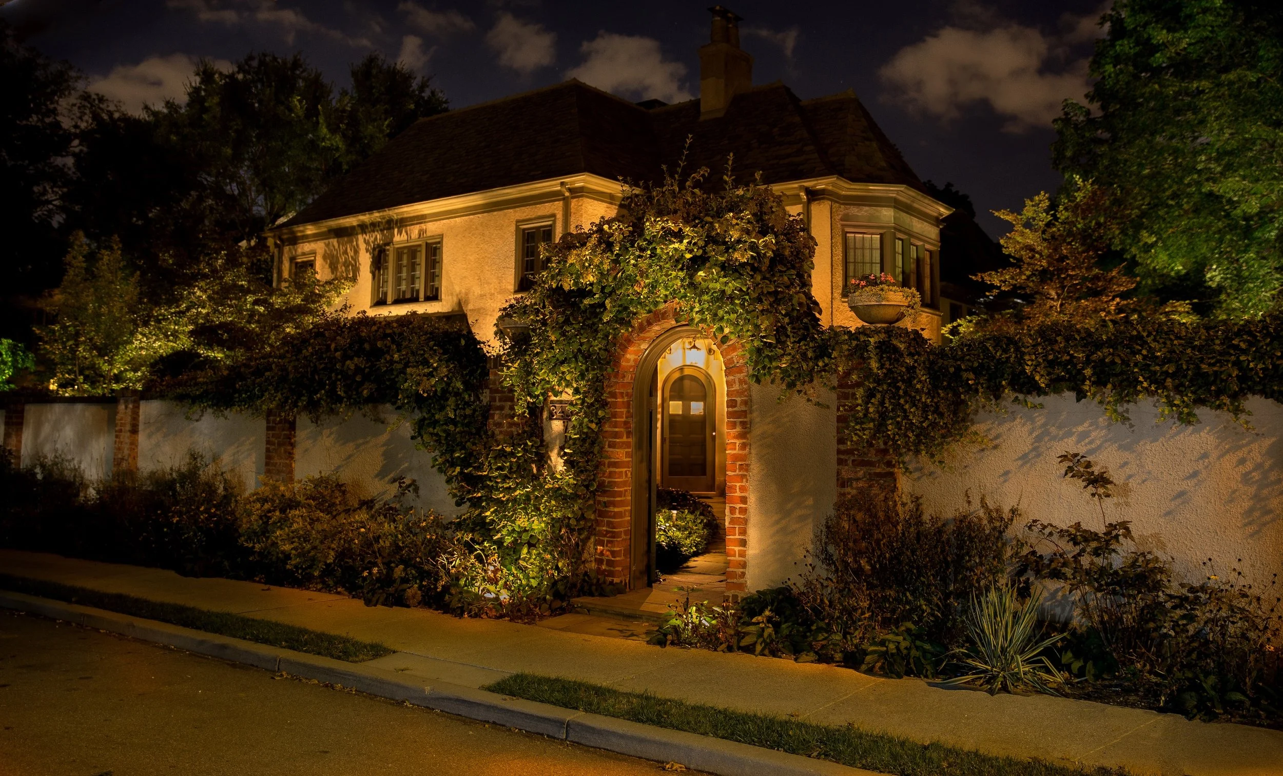 A house at night with warm exterior lighting, surrounded by trees and a garden, with a brick archway entrance and a sidewalk in front.