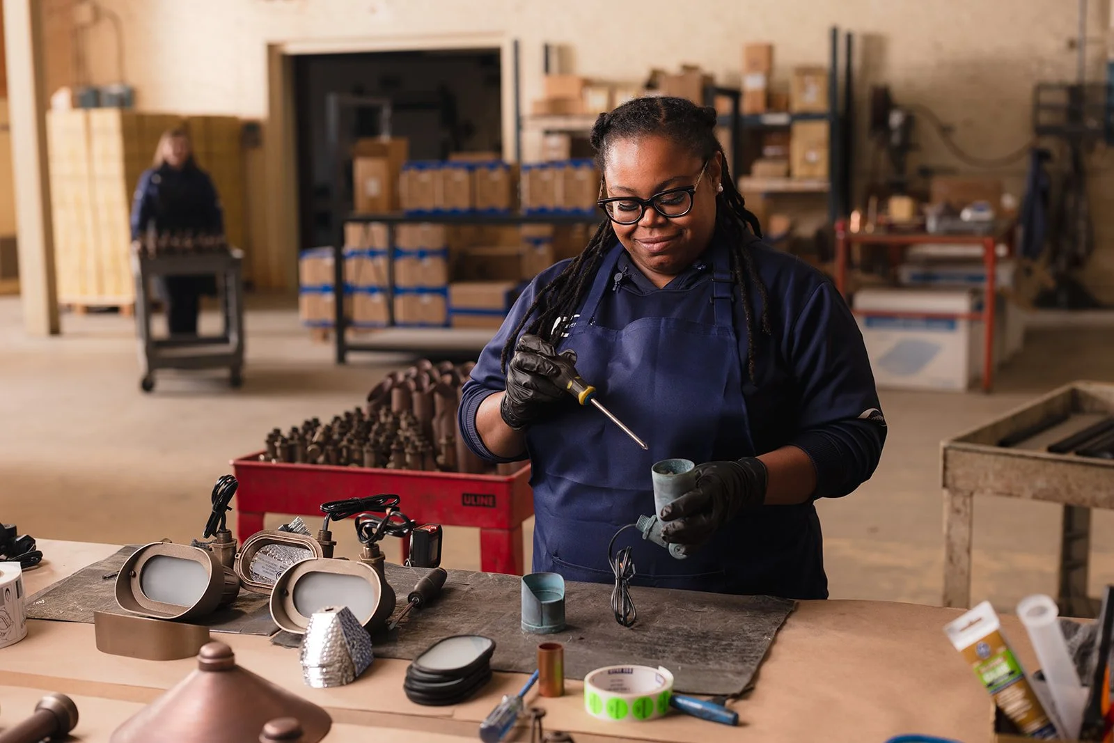 A woman in safety glasses and gloves works with small metal components in a workshop filled with tools and parts.