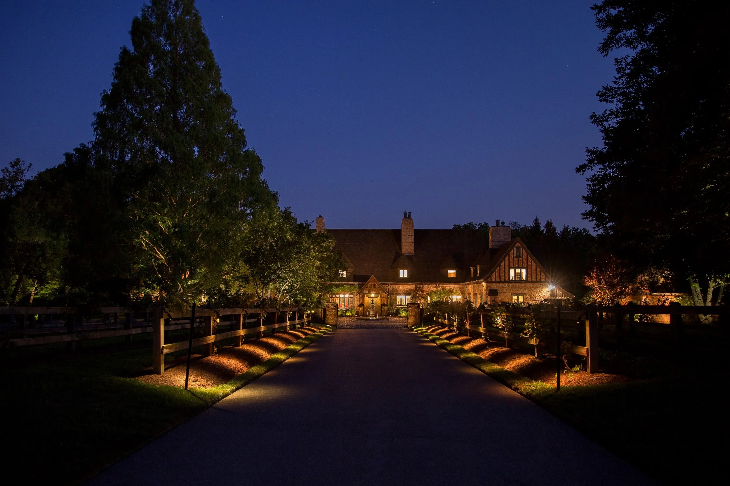 Nighttime scene of a large house with lit windows, surrounded by trees, with a starry sky overhead and a wooden fence lining the driveway.