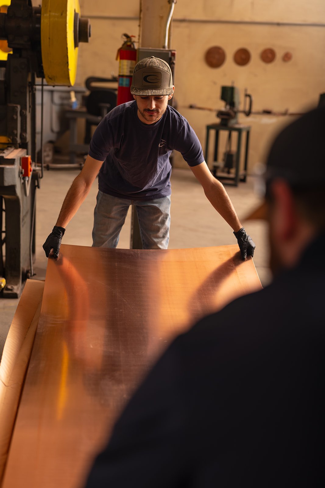 Two men in a workshop handling a large sheet of copper, one man bending down and the other man in the background.