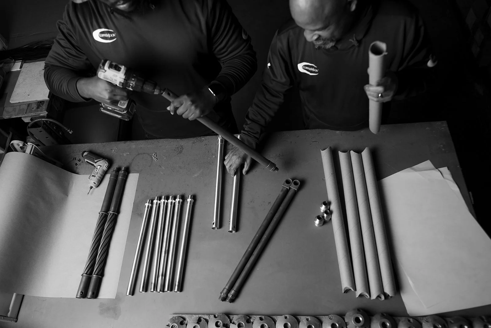 Two men working with pipes and tools on a workbench. One is holding a drill and the other is holding a cylindrical pipe. Various pipes and fittings are laid out on the table.