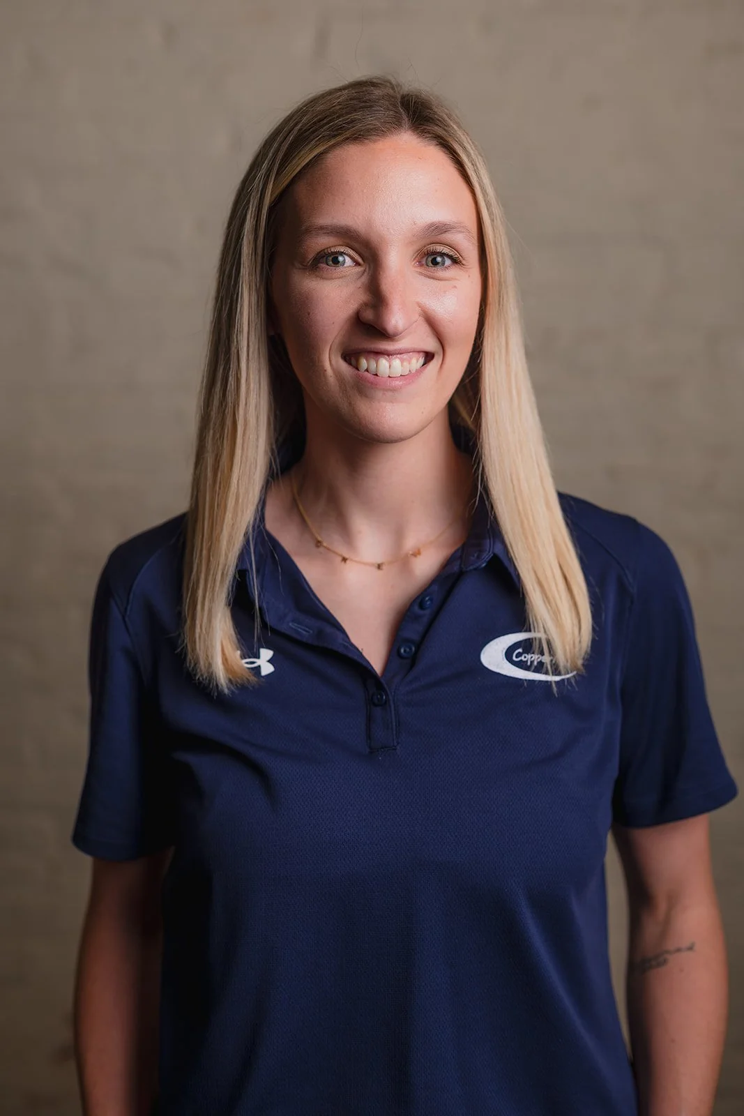 A smiling woman with long blonde hair wearing a navy blue polo shirt with logos, standing in front of a beige wall.