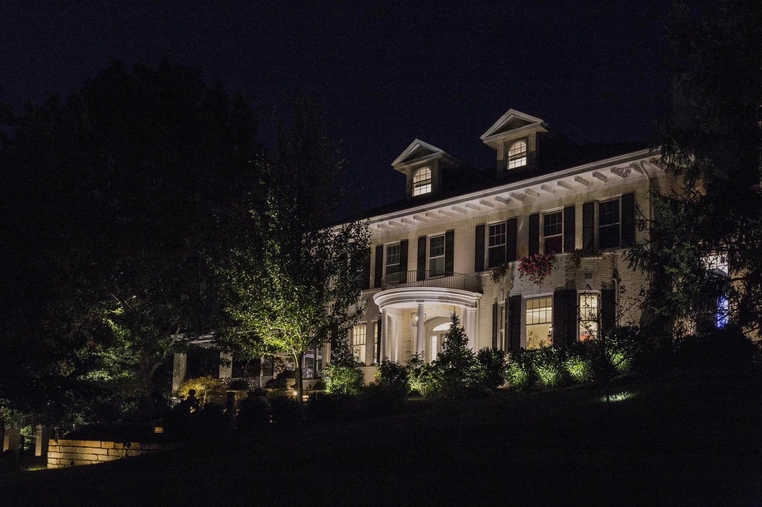 Nighttime view of a well-lit multi-story house with a prominent entrance, surrounded by trees and shrubbery.