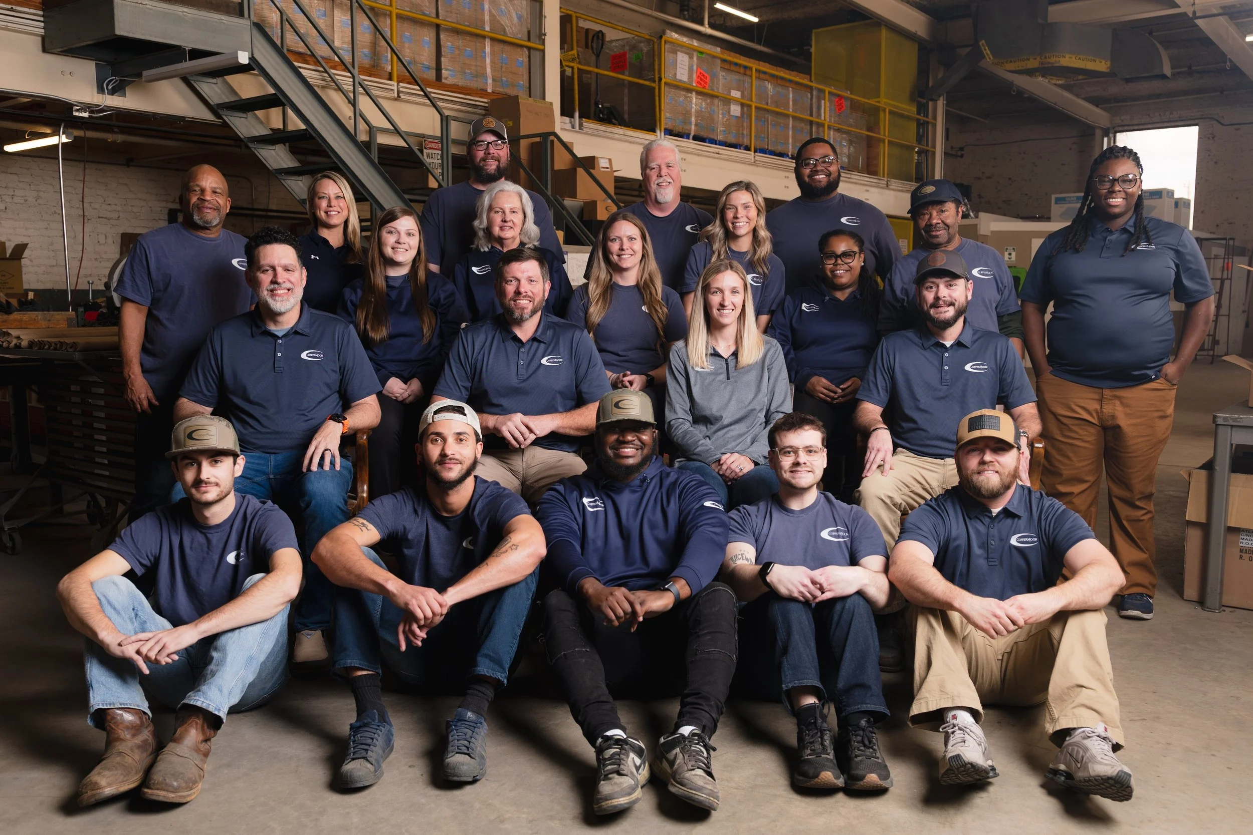 Group of people posing for a photo inside a warehouse. The group consists of men and women wearing matching blue and gray company shirts. Some are sitting on the floor, some are standing behind, and others are standing on a platform. The warehouse has pallets and boxes in the background.