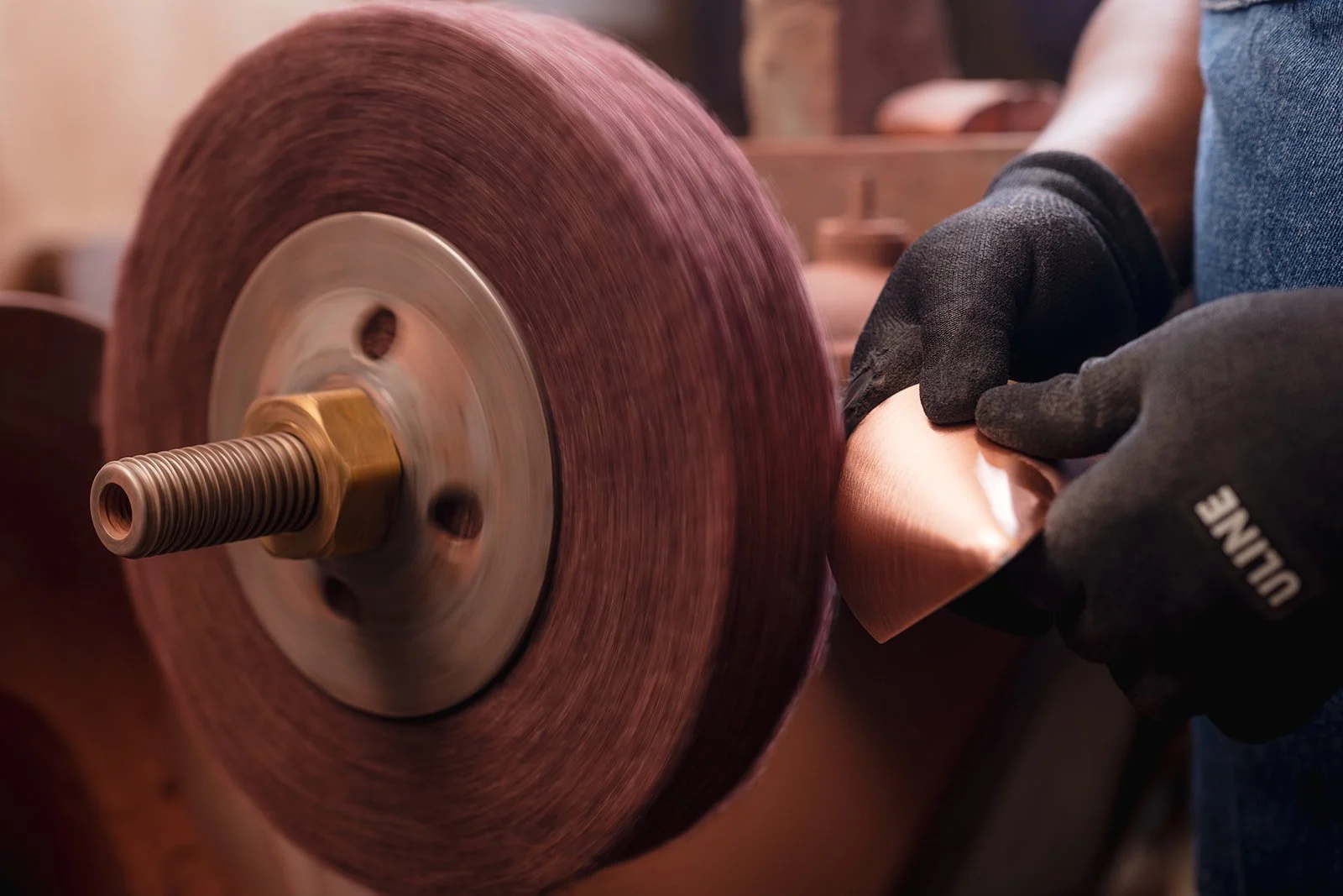 A person wearing black gloves handling a copper sheet near a large industrial grinding wheel.