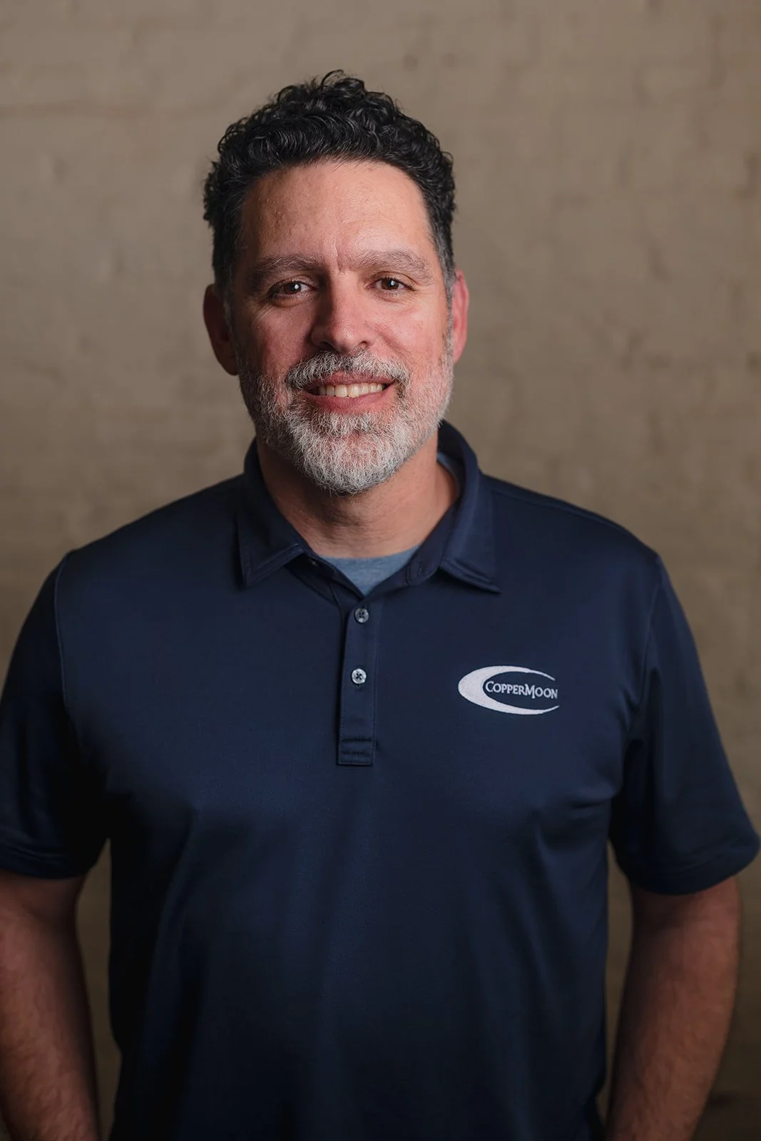 A smiling man with dark curly hair and a beard, wearing a navy polo shirt with a 'CopperMoon' logo, standing in front of a beige textured wall.