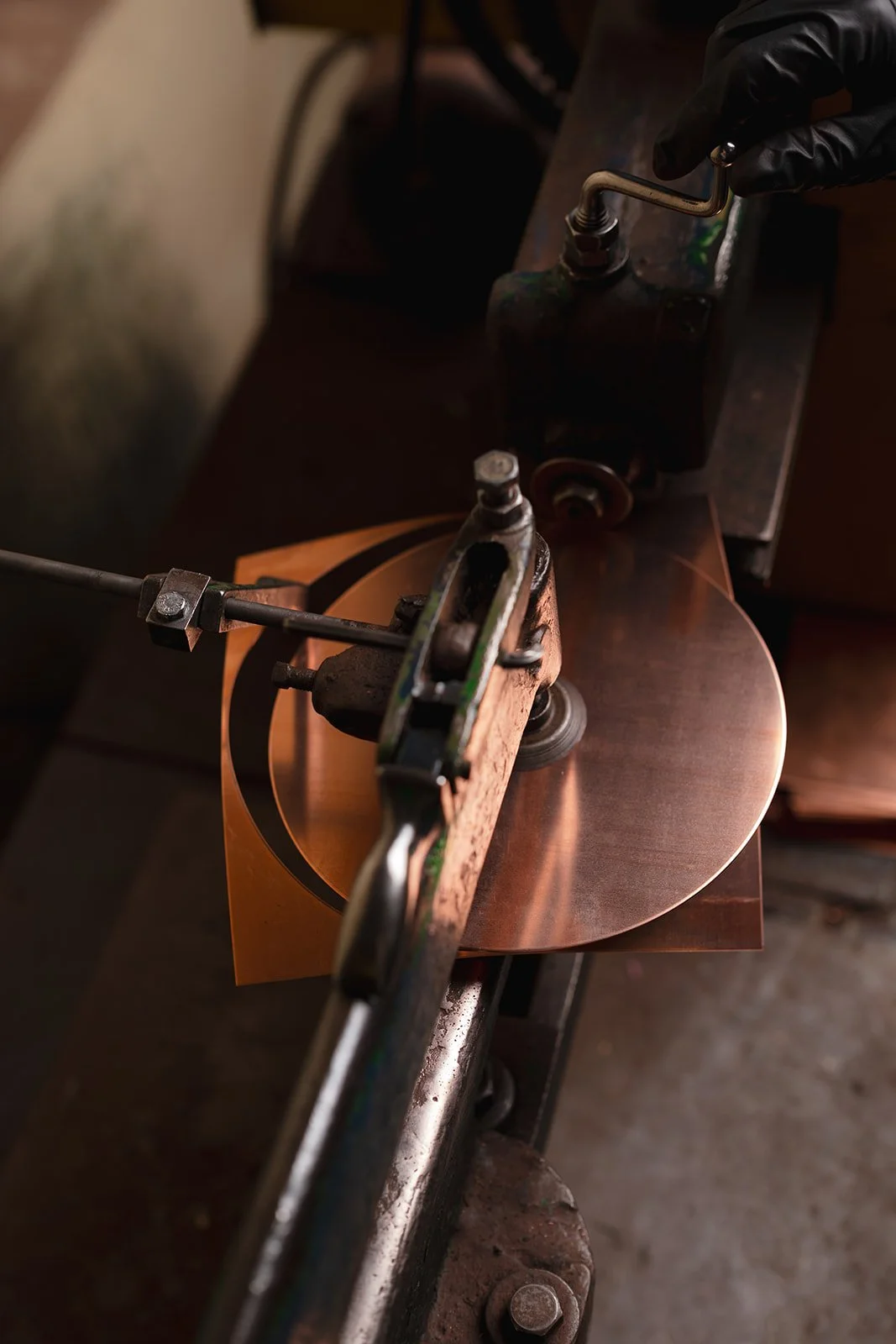 A close-up of a mechanical lathe machine shaping a copper disc, with a black-gloved hand operating the machine.