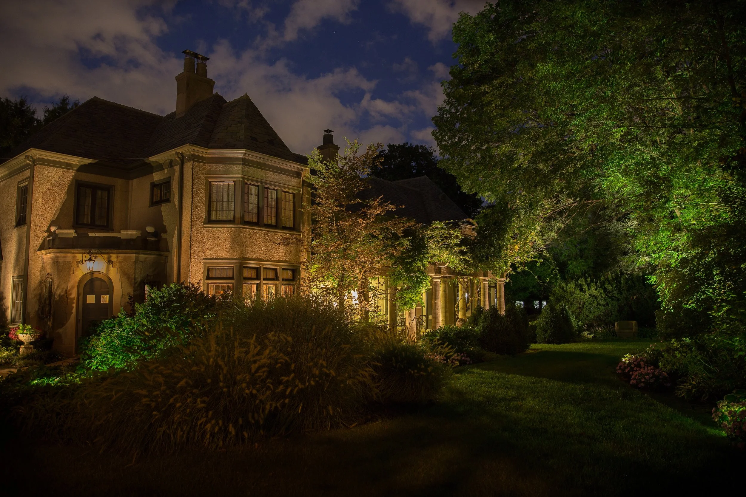 A large, elegant house at night with exterior lights illuminating the garden and trees in the yard.