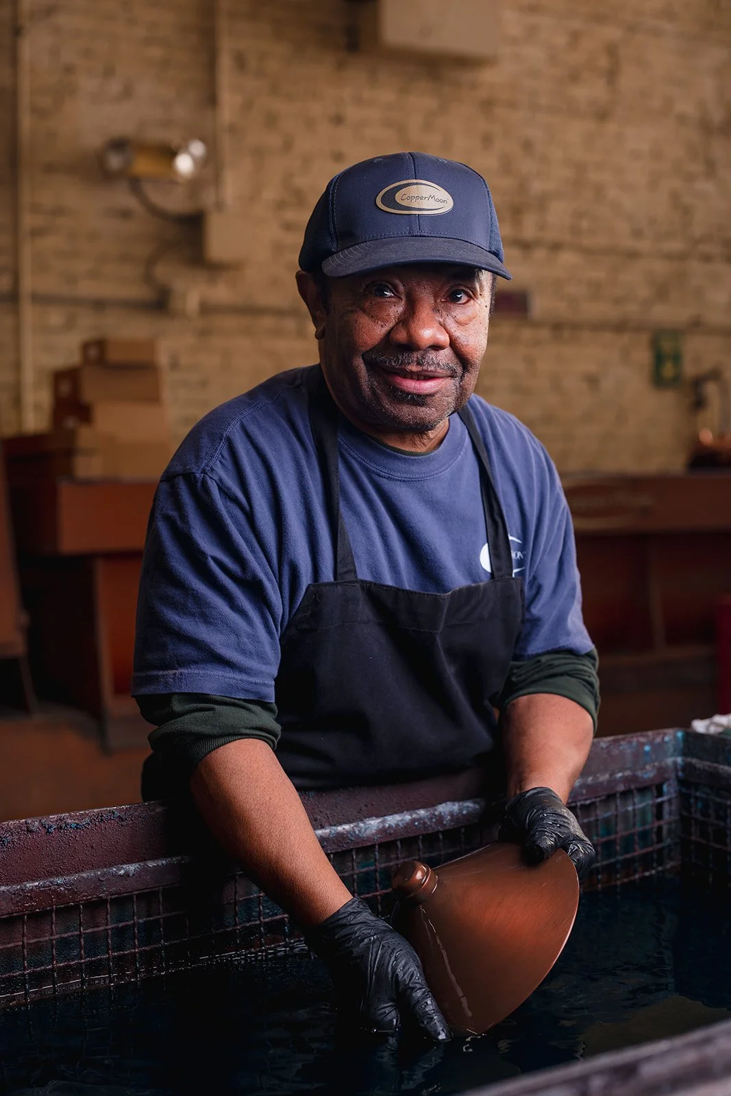 A man wearing a navy blue cap, blue T-shirt, and black apron, standing in front of a large water basin, holding a guitar-shaped object with black gloves, inside a rustic indoor setting with a brick wall in the background.