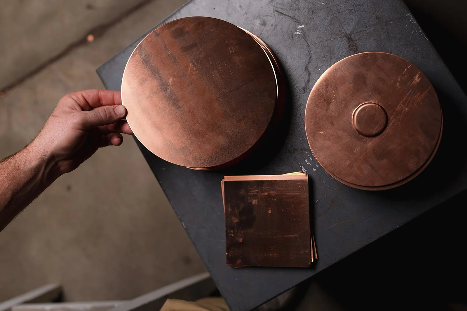 Person holding a round copper metal piece, with two other copper round pieces and a smaller copper rectangle on a black work surface.