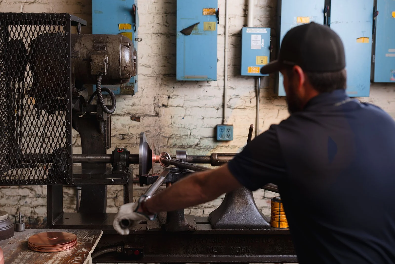 A man wearing a black cap and dark shirt working with a metal lathe machine in a workshop, shaping a metal rod.