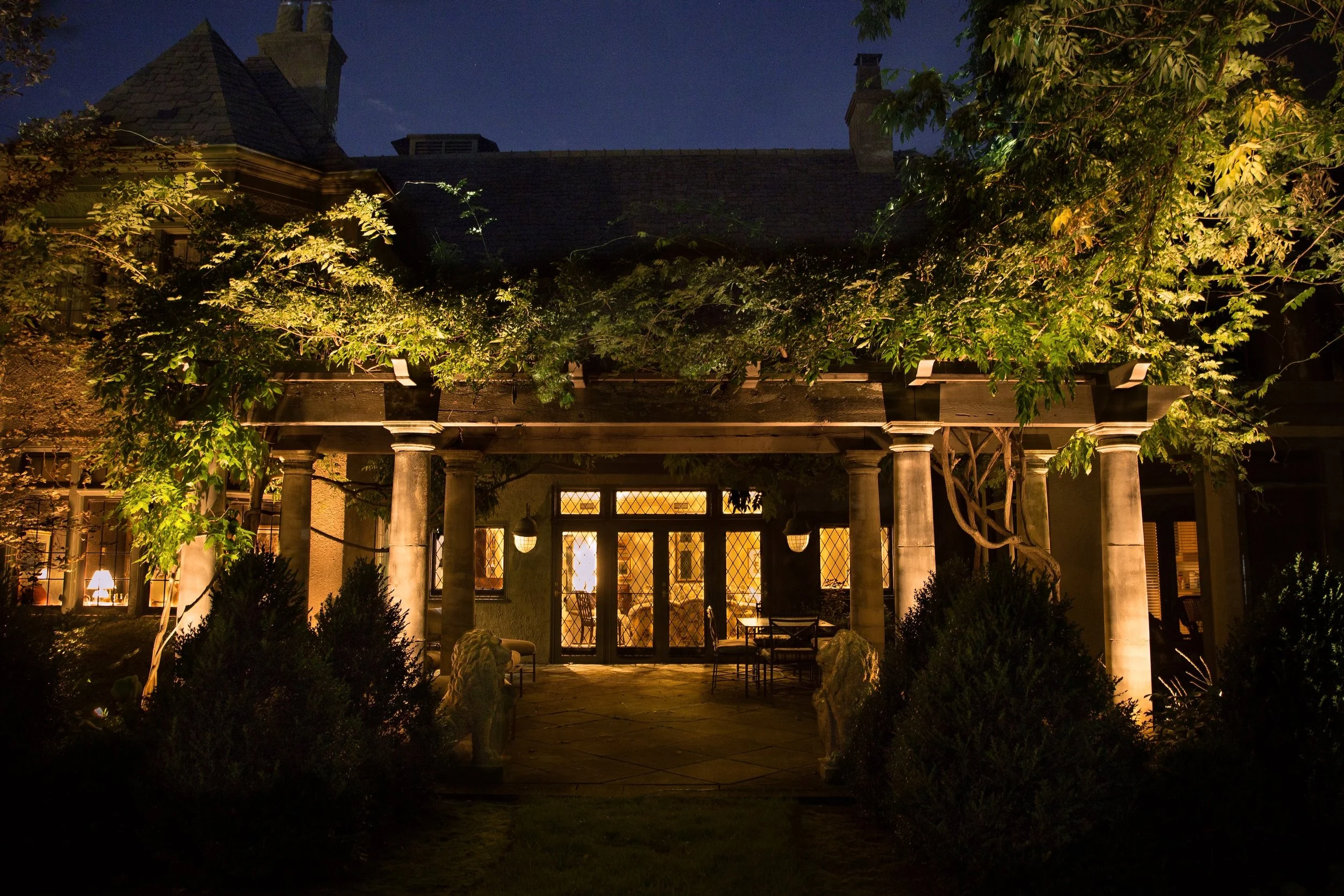 A house with a yard at night, illuminated by exterior lights. The house has a sloped roof, windows, and columns supporting a porch or balcony. Trees and bushes surround the house, with some lighting on the foliage.