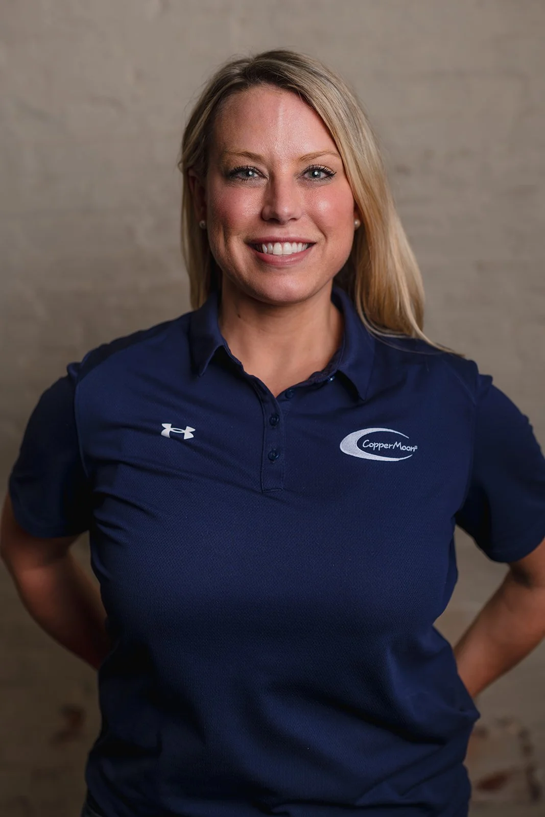 A woman with blonde hair in a blue polo shirt smiling with her hands on her hips against a beige background.