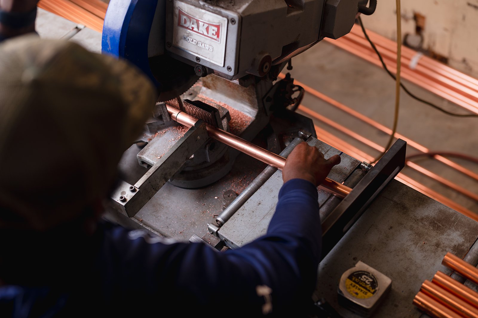 A worker using a band saw to cut a copper pipe in a workshop, with copper pipes on the work surface.