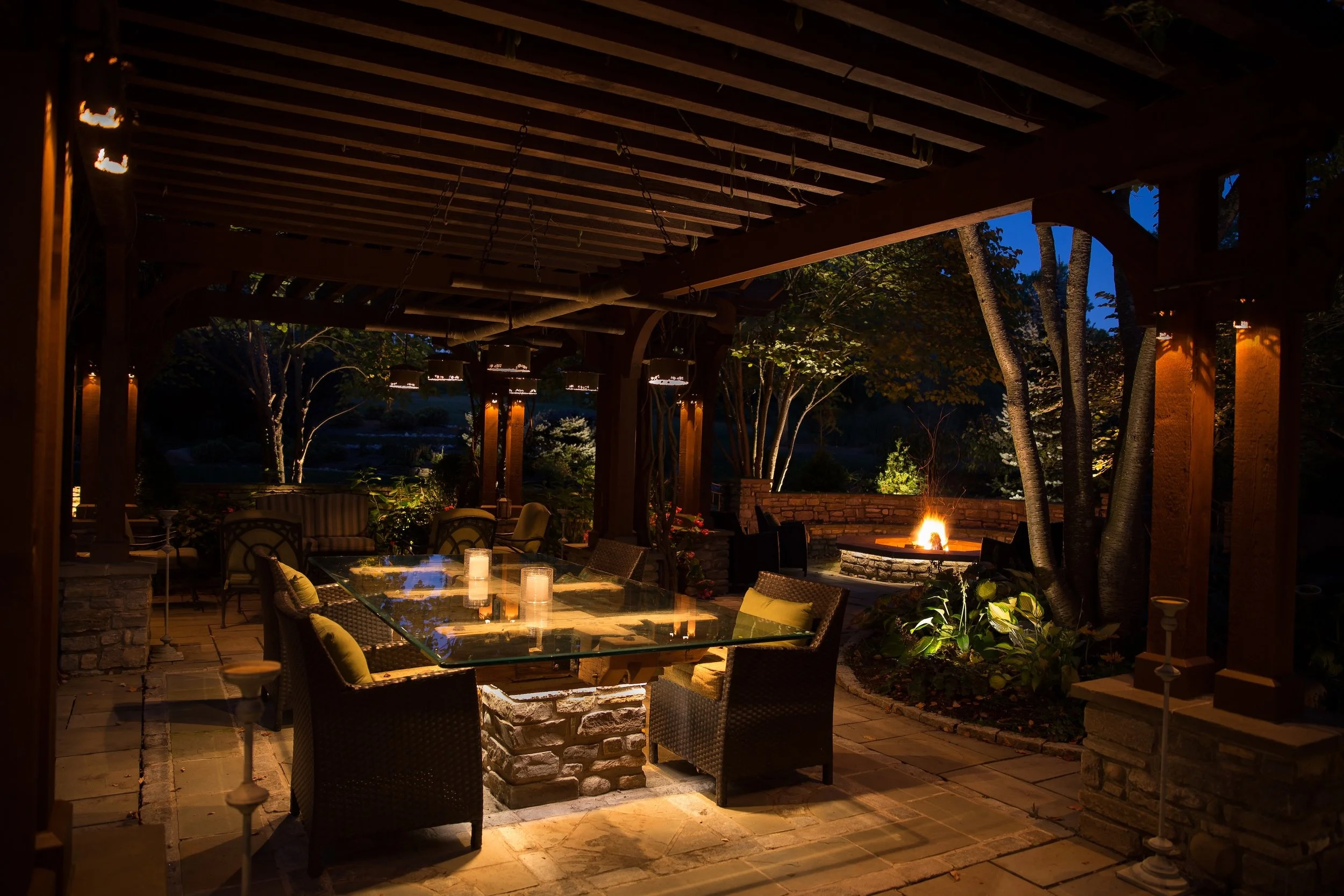 Outdoor patio at night with a glass table, chairs, a fire pit, string lights, and trees in the background.