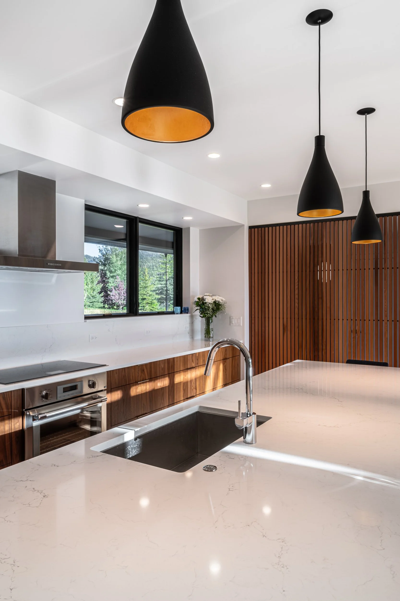 Modern kitchen with white marble countertops, black pendant lights, wooden cabinets, stainless steel appliances, and a large window with a scenic view