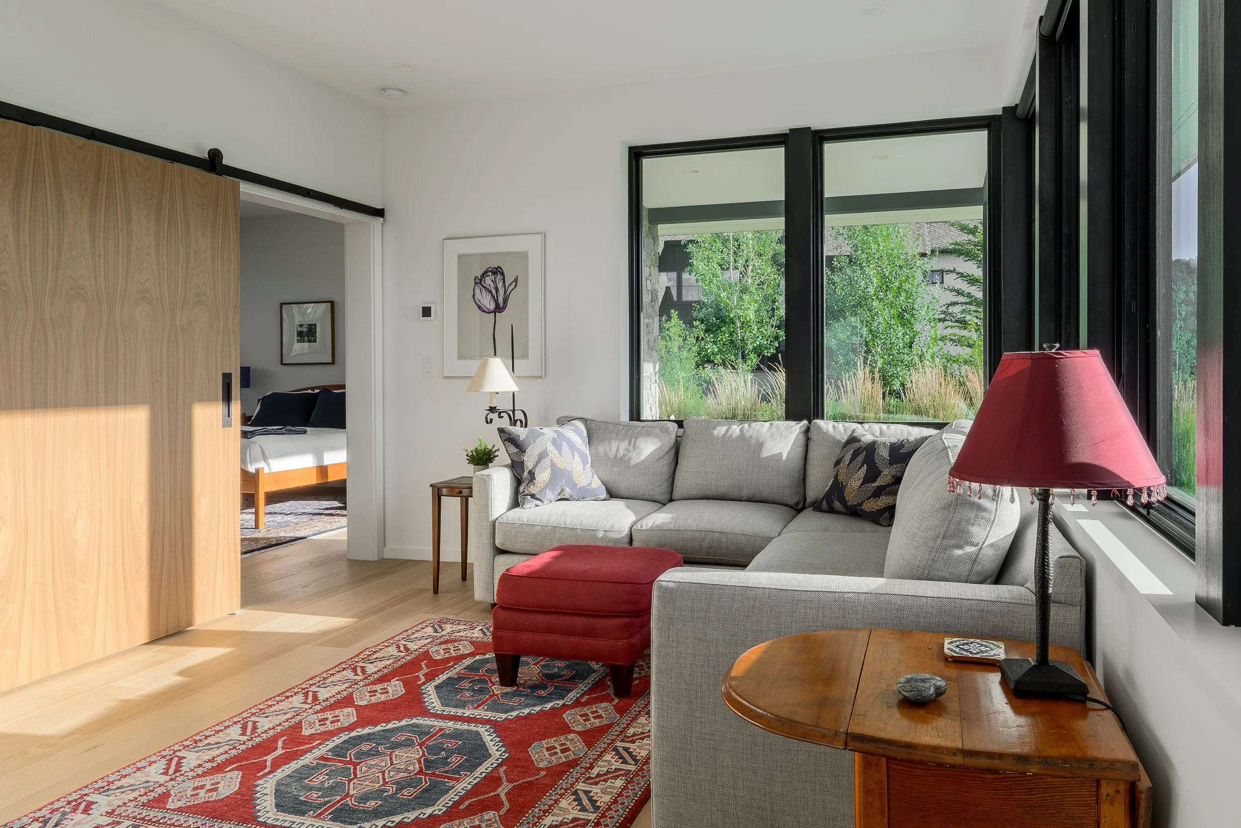 Living room with a light gray sectional sofa, red ottoman, and a red lampshade on a wooden side table, large windows with a view of green trees, and a sliding wooden door leading to a bedroom.