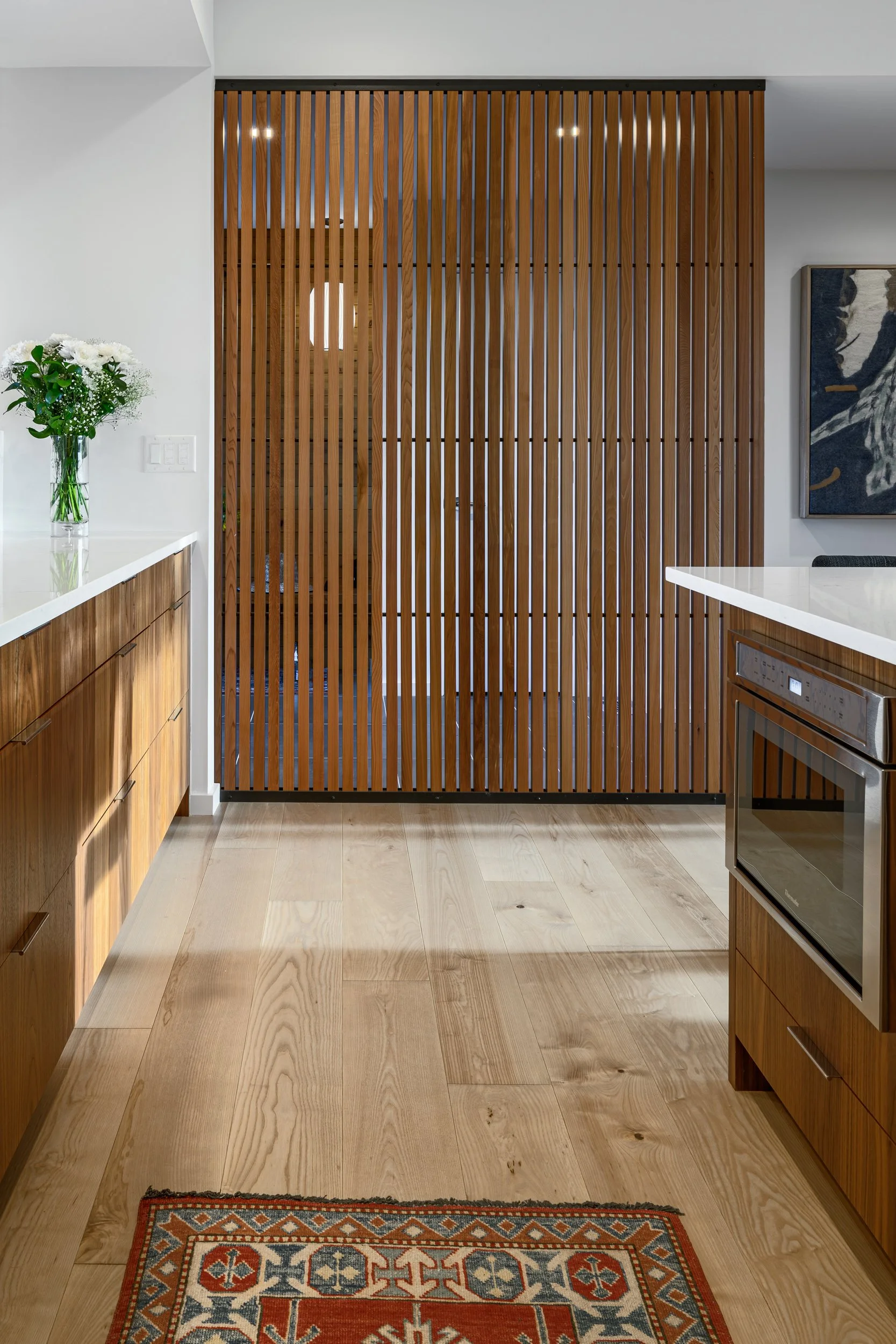 A modern kitchen with wooden cabinets, white countertops, a patterned area rug, a flower vase on the counter, and a partition made of vertical wooden slats.