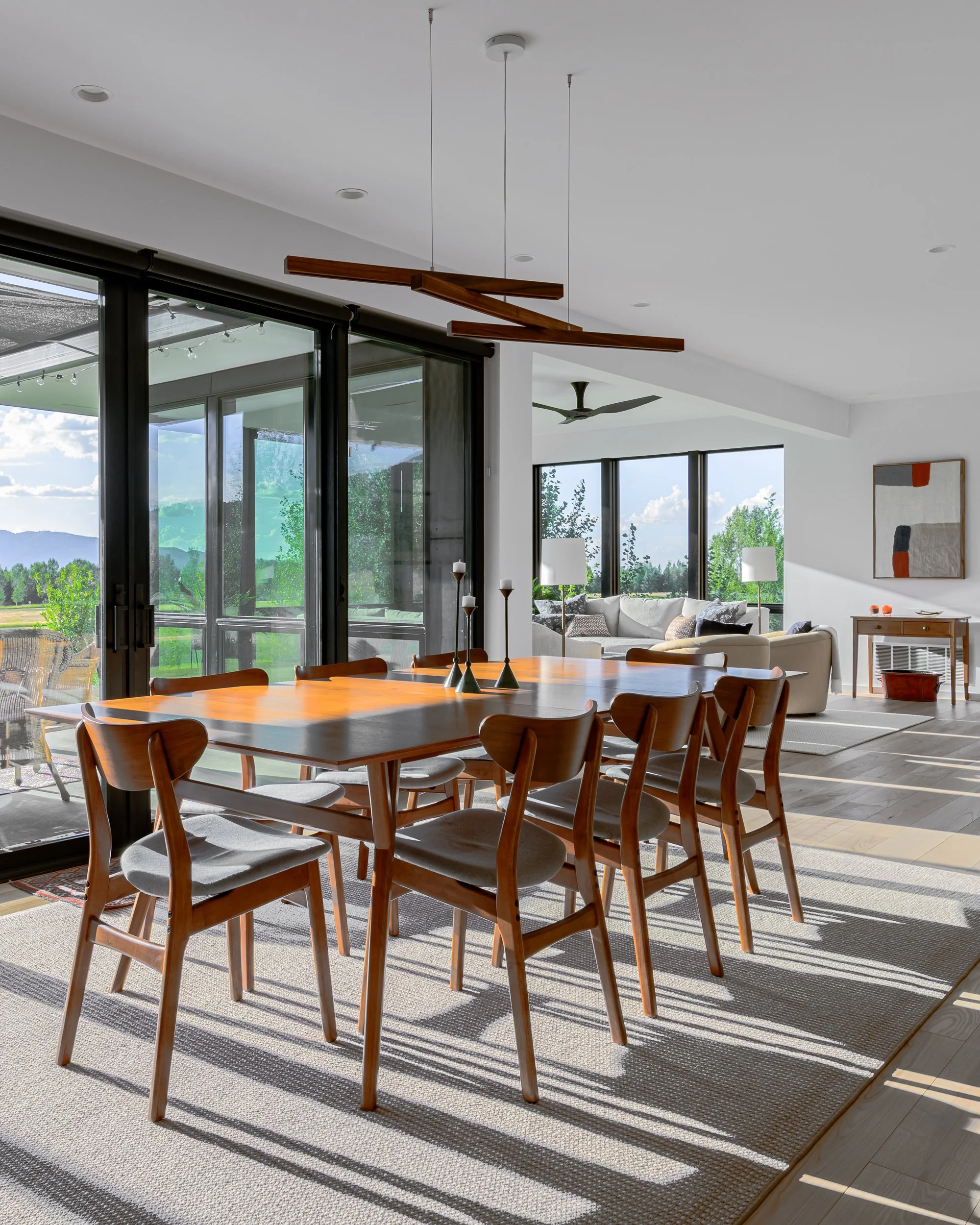 Modern dining room with wood dining table, eight chairs, and a living area with beige sofas, large windows, and outdoor greenery.